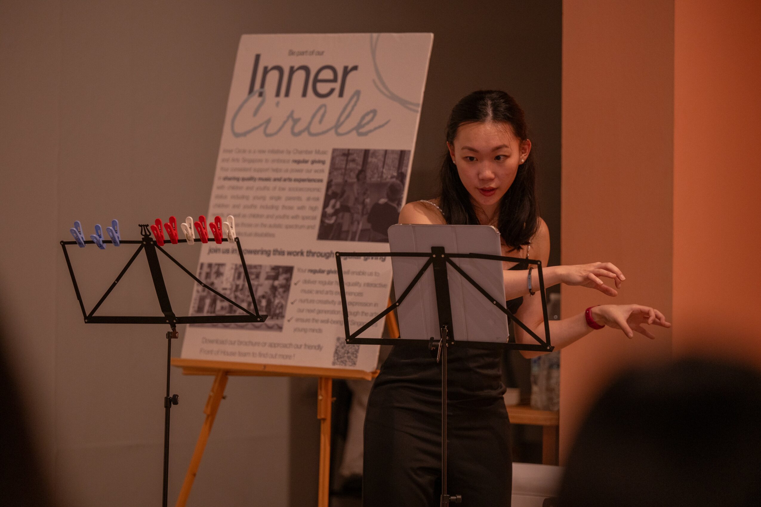 Young woman standing in front of a poster, gesturing with her hands, with music stands nearby.