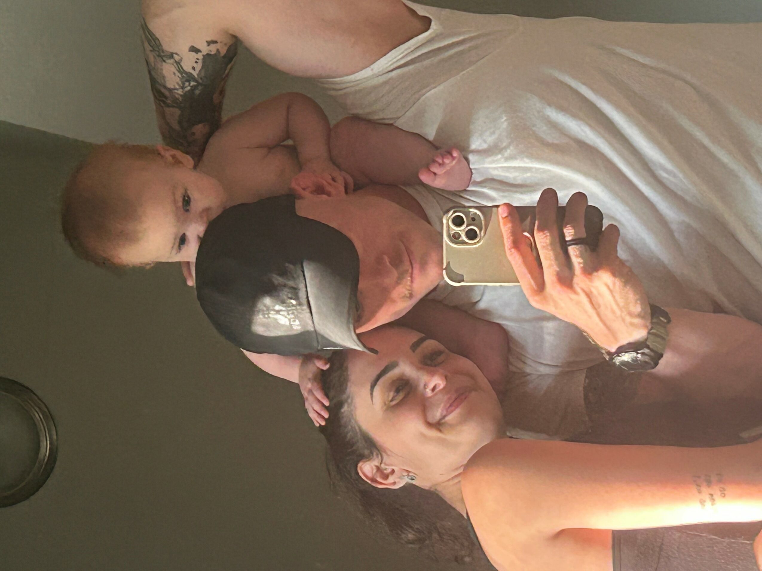 Person lying down holding a baby, taking a selfie with a smartphone, both smiling, in a room with a plain background.