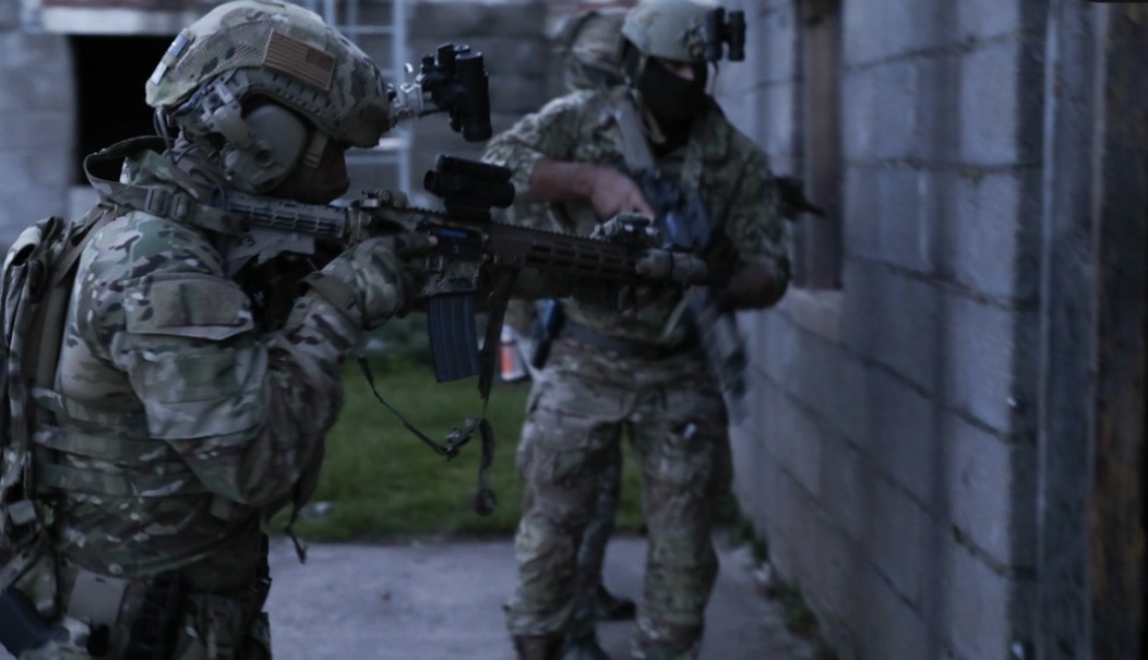 Two soldiers in camouflage gear aiming rifles beside a wall at dusk.
