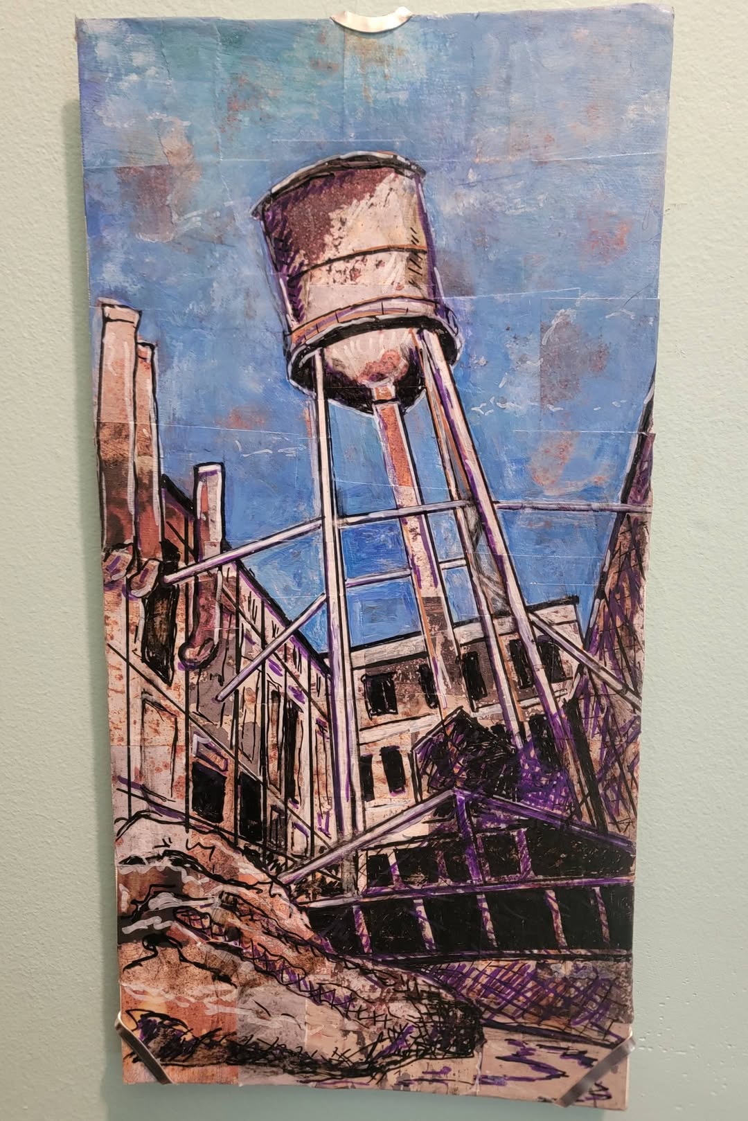 Water tower tilted on a building with a blue sky background, surrounded by structures and a rocky foreground.