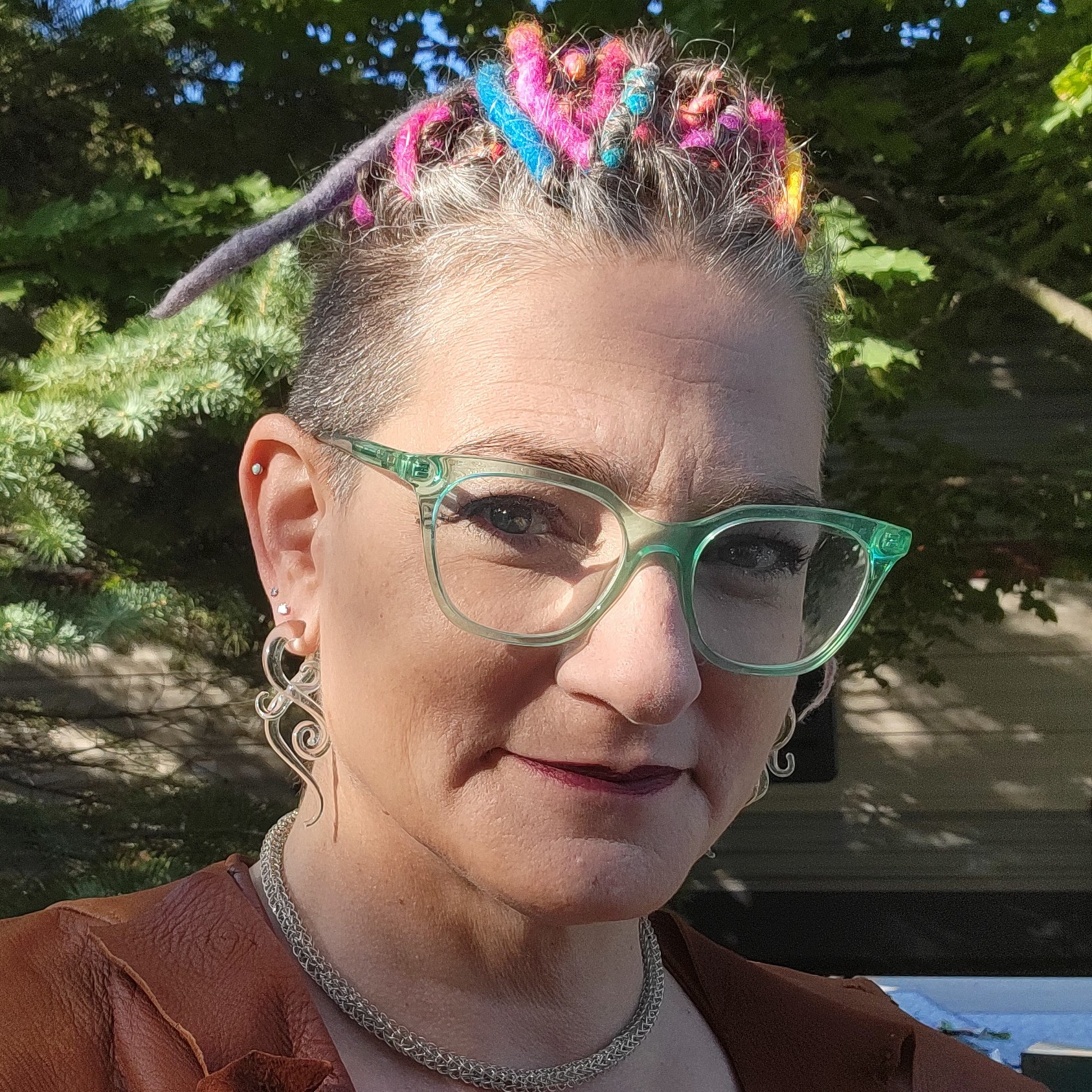 Woman with colorful dreadlocks, glasses, and earrings outdoors, smiling slightly, with trees in background.