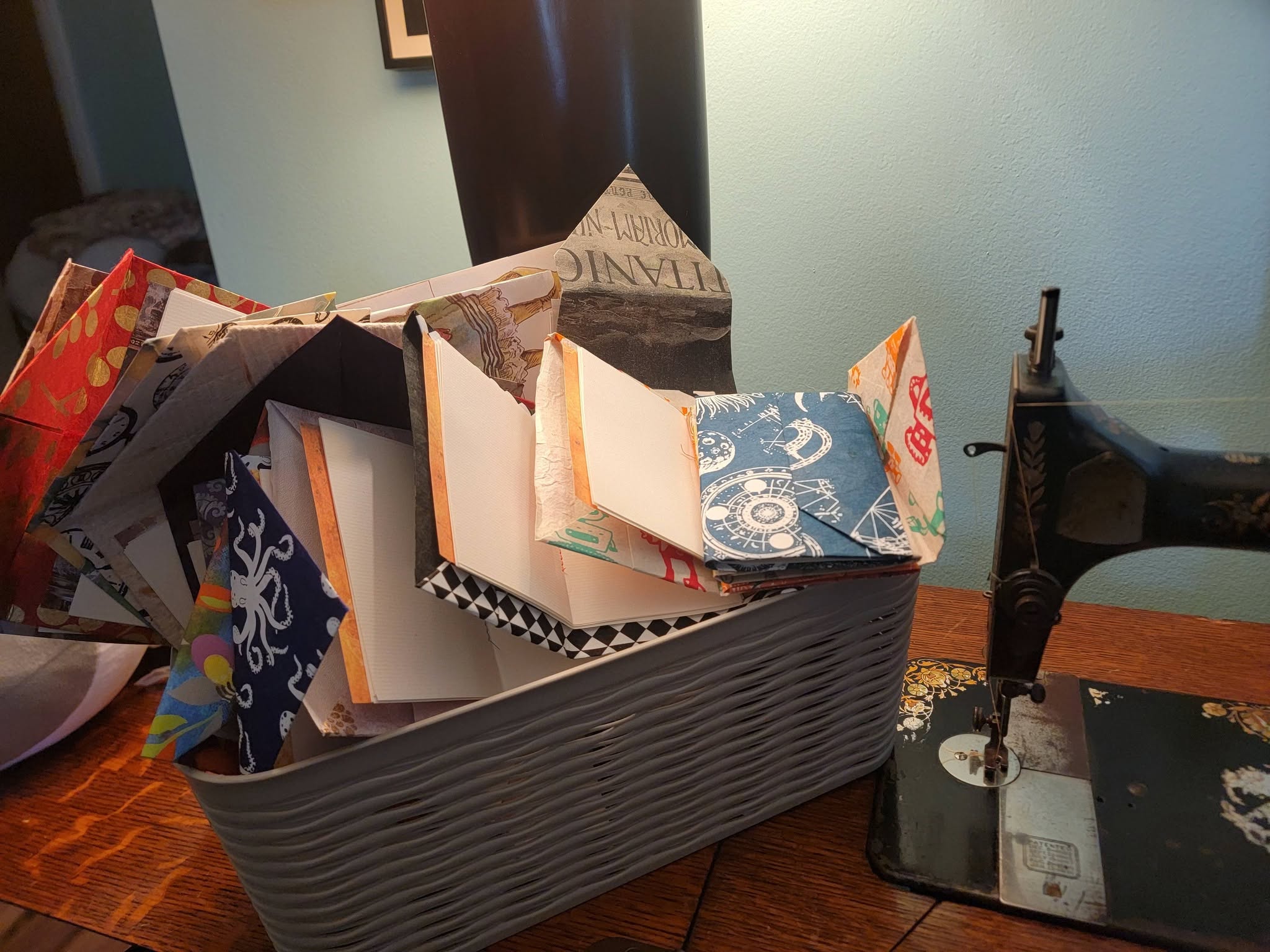 A gray rectangular container filled with colorful, patterned paper and cards on a wooden table, with a sewing machine nearby.