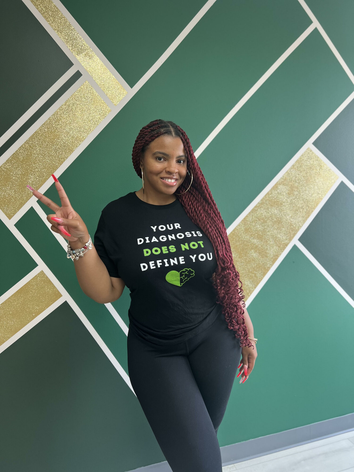 Smiling woman with long braided hair making a peace sign, standing against a geometric green and gold wall.
