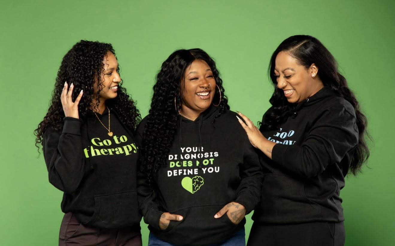 Three women smiling and laughing together against a green background, wearing black hoodies with text. One woman touches another's shoulder.