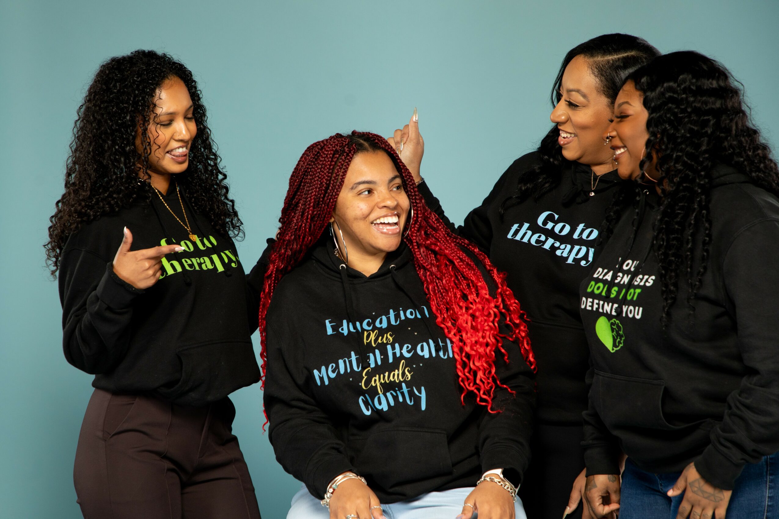 Four women smiling and talking, wearing black hoodies with colorful text, against a light blue background.