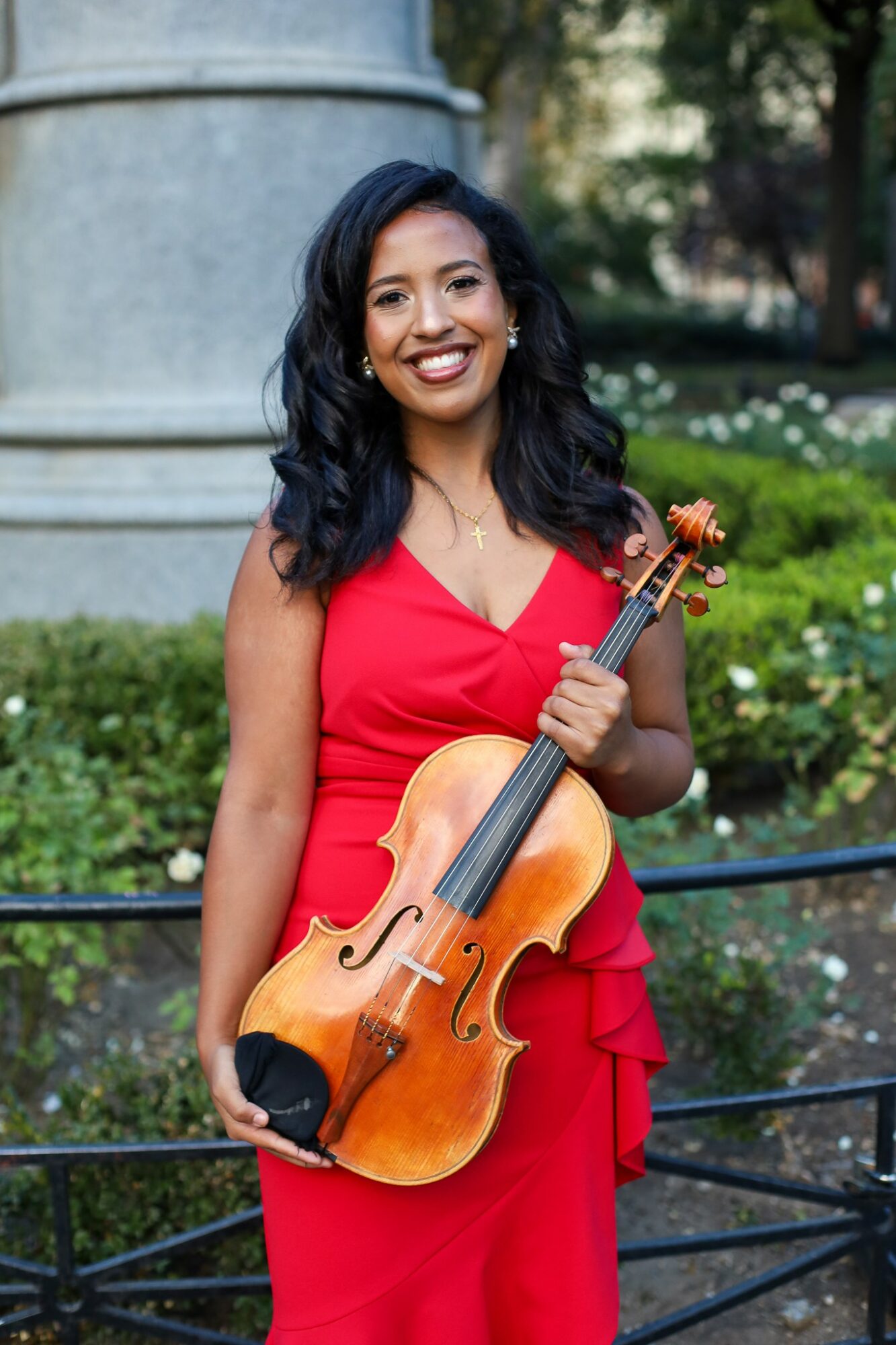 Woman in red dress holding a violin outdoors, smiling, with greenery and a stone structure in background.