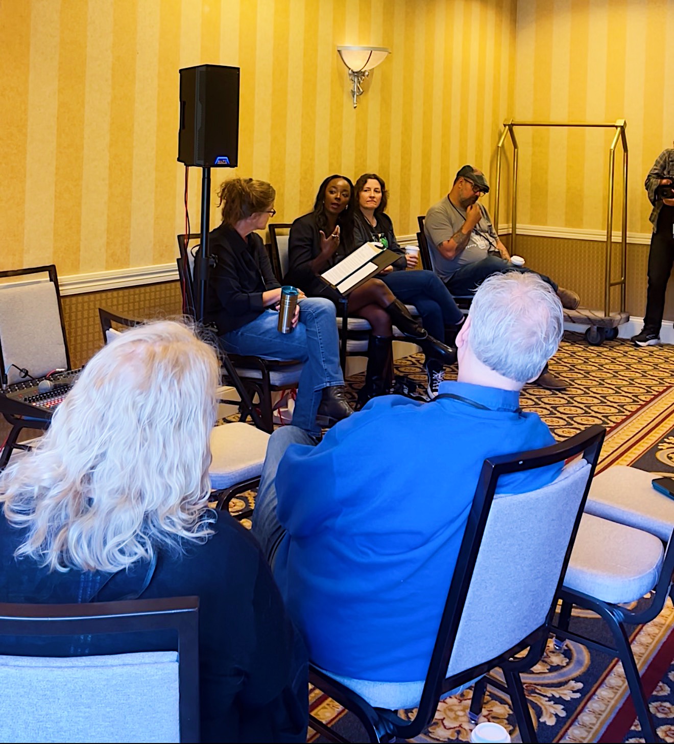 Four people seated on chairs in a panel discussion, with an audience in a yellow-walled room, speaker equipment, and a patterned carpet.