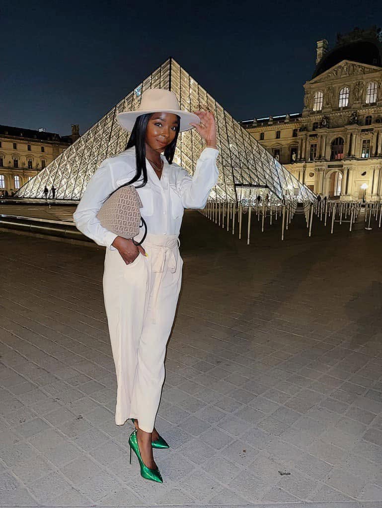 Woman in white outfit and green heels standing in front of a glass pyramid at night.