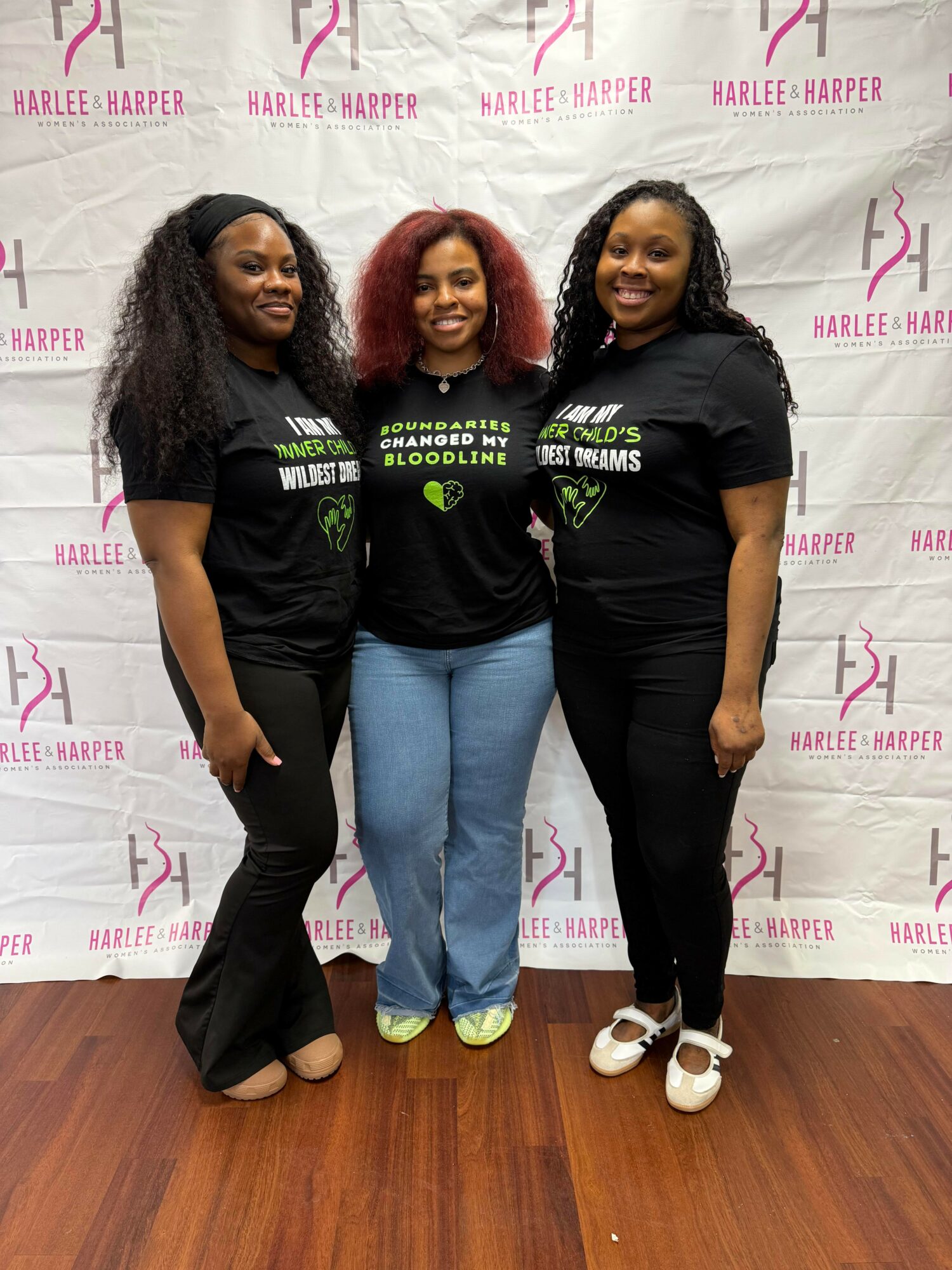Three women stand together in front of a Harlee Harper backdrop, smiling, wearing black T-shirts, and jeans or black pants.