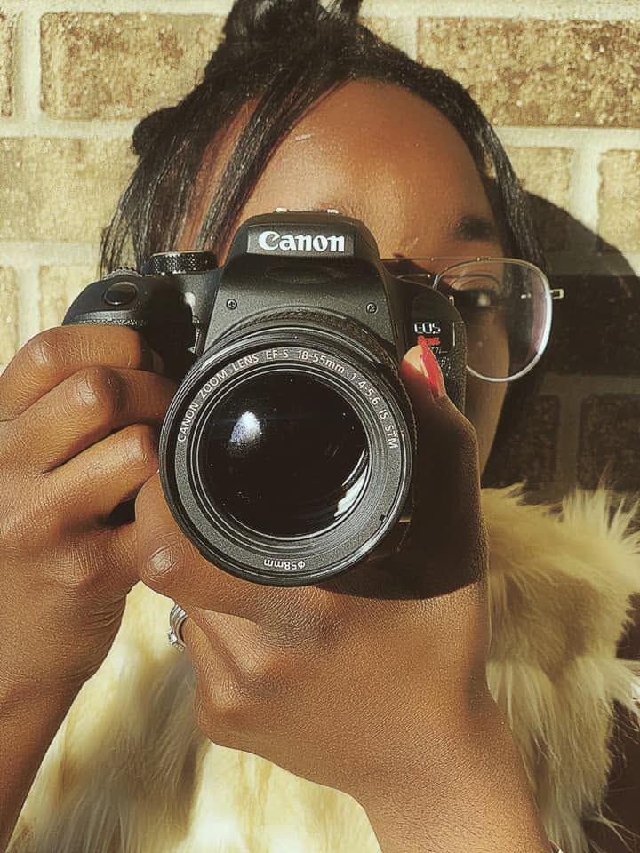 Person holding a Canon camera in front of face, with glasses and a fur coat, standing outdoors against a brick wall.