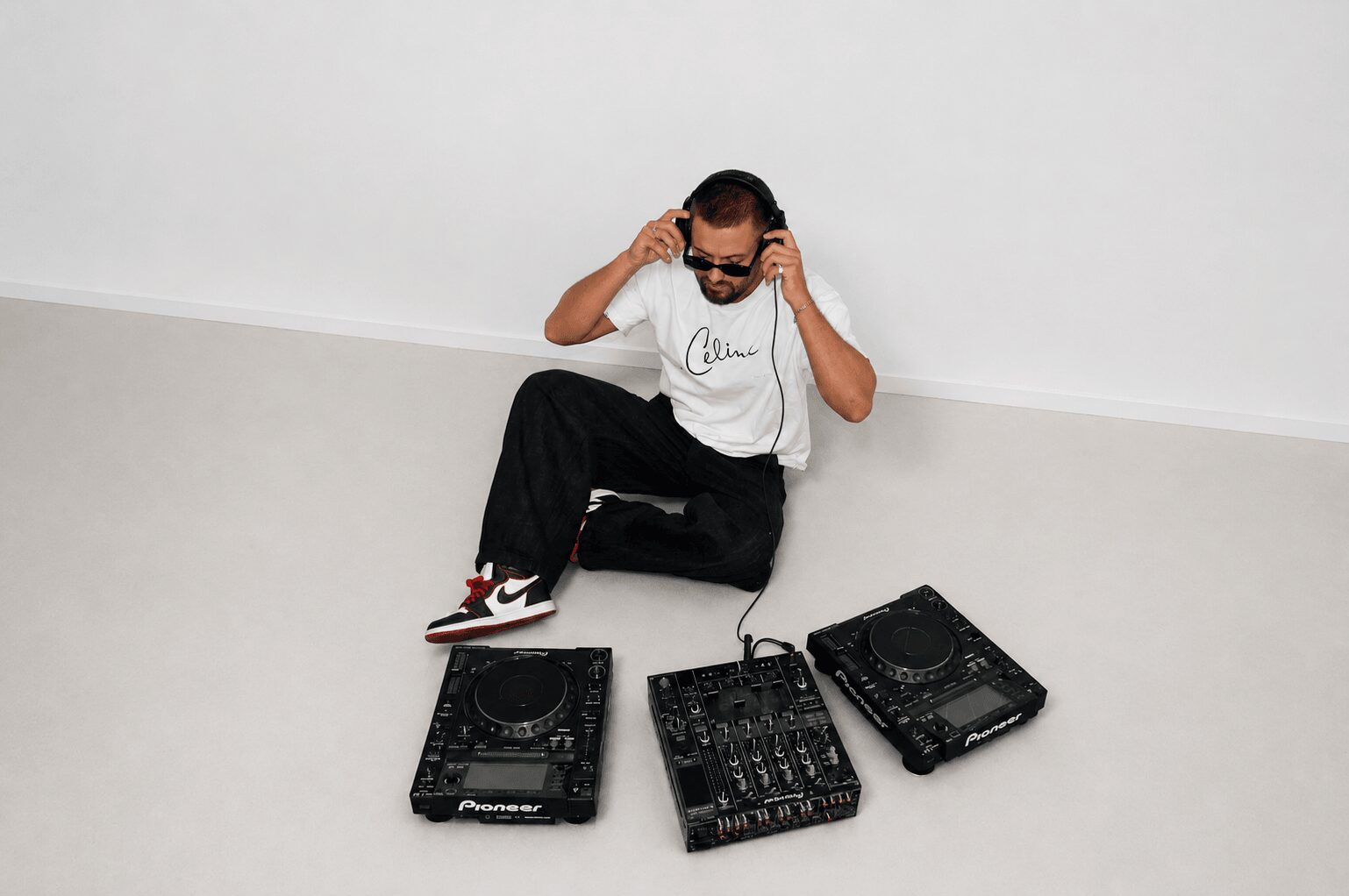 Person with headphones sitting on the floor with DJ equipment, white background, casual clothing, sunglasses.