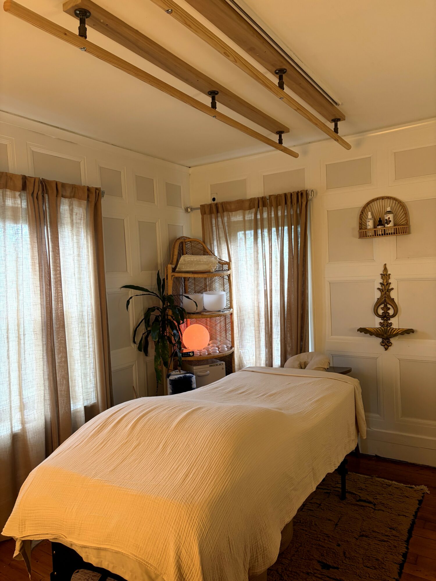 A bedroom with a bed, curtains, a plant, and wall decor, illuminated by natural light from windows.