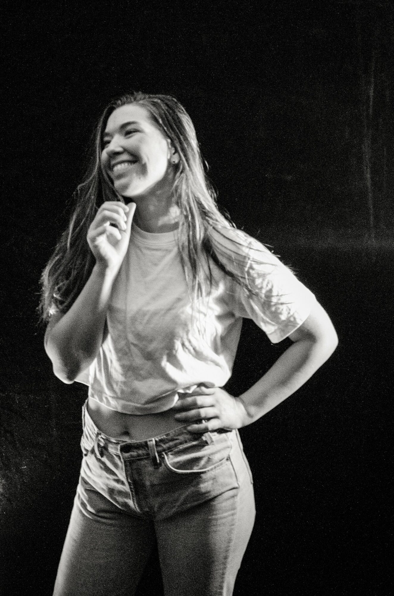 Smiling woman with long hair, wearing a t-shirt and jeans, standing with hand on hip against dark background.