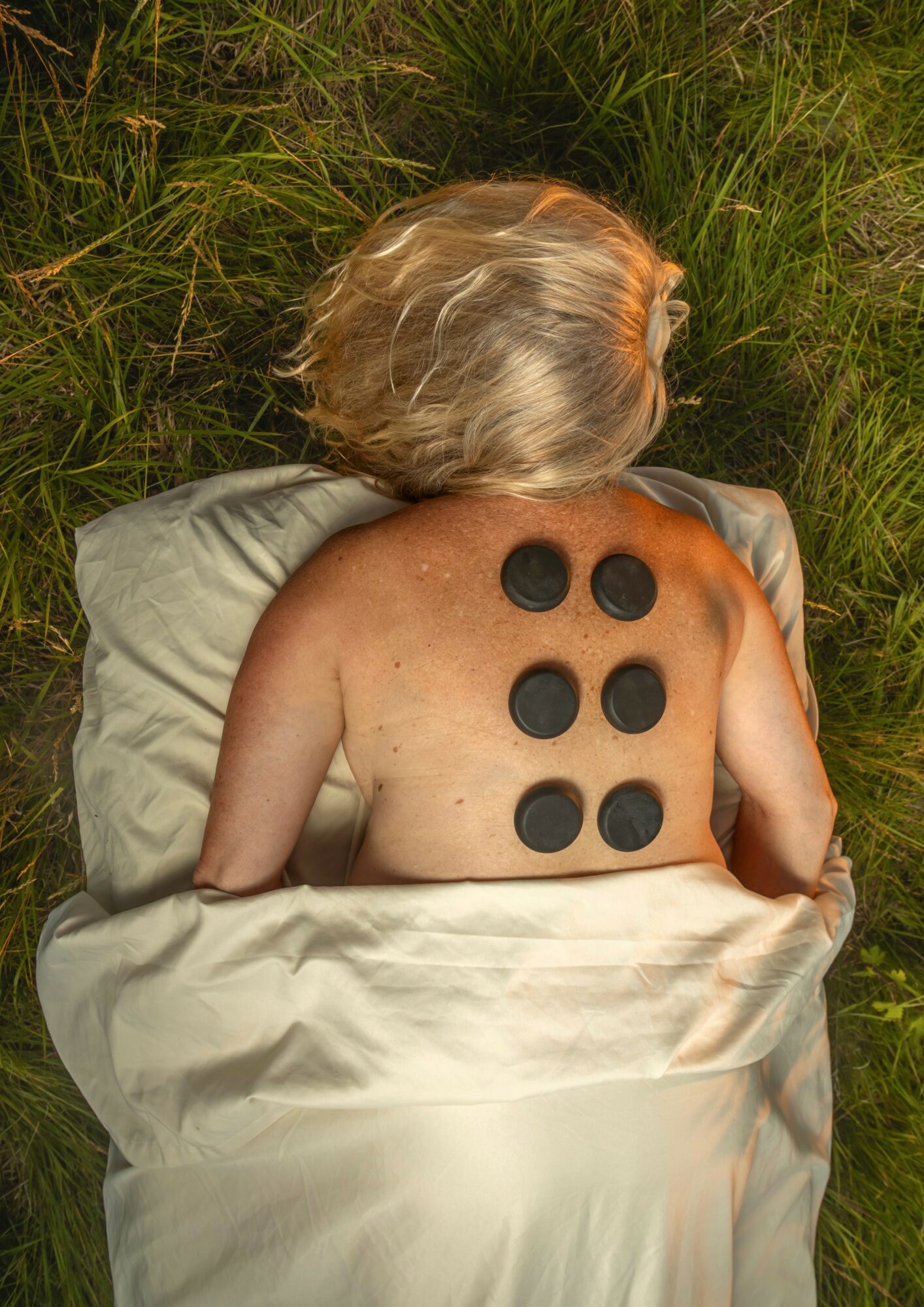 Person lying on grass with six black circular objects on their back, head turned to the side, surrounded by green grass.