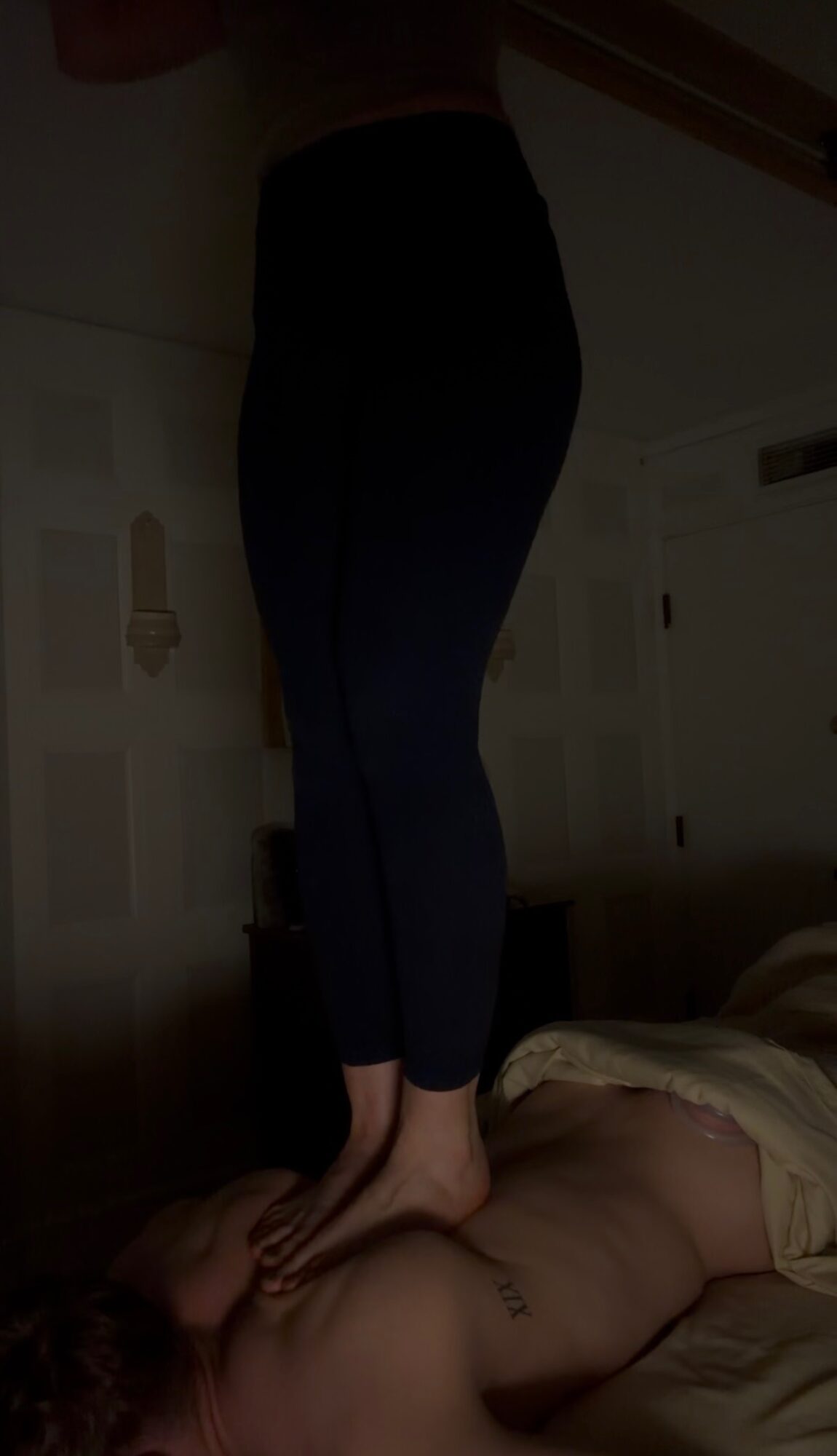 Person standing on a bed with a person lying down, in a dimly lit room.