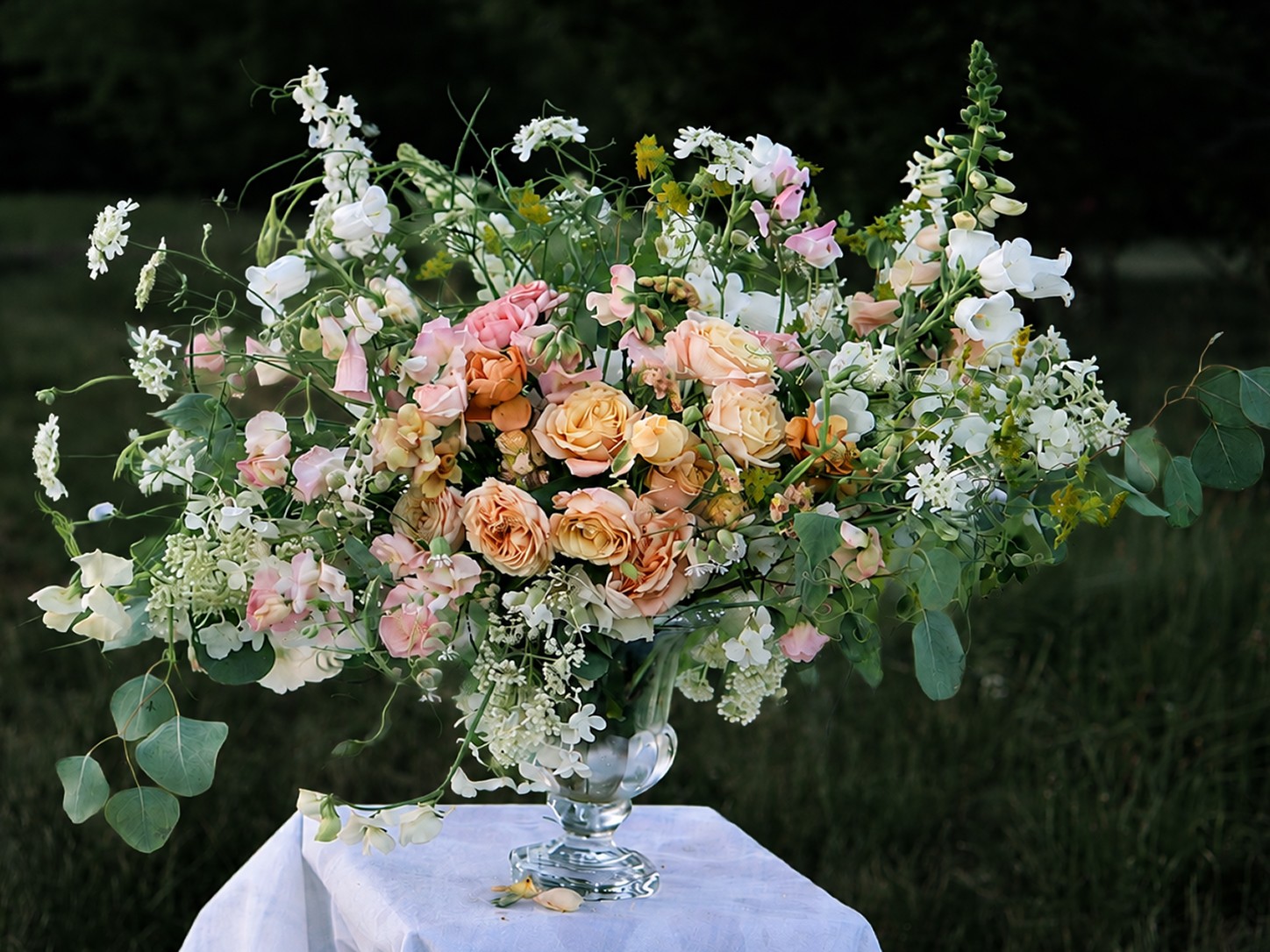 Large floral arrangement with pink, peach, and white flowers, green leaves, in a glass vase on a white cloth outdoors.
