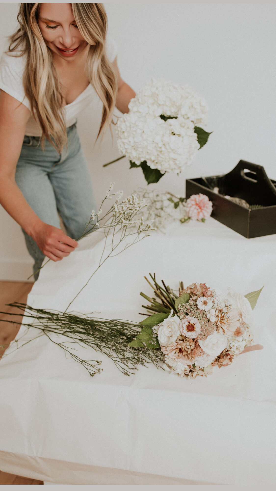 Person arranging flowers on a table with a box of flowers nearby, smiling, with white and pink flowers, and greenery.