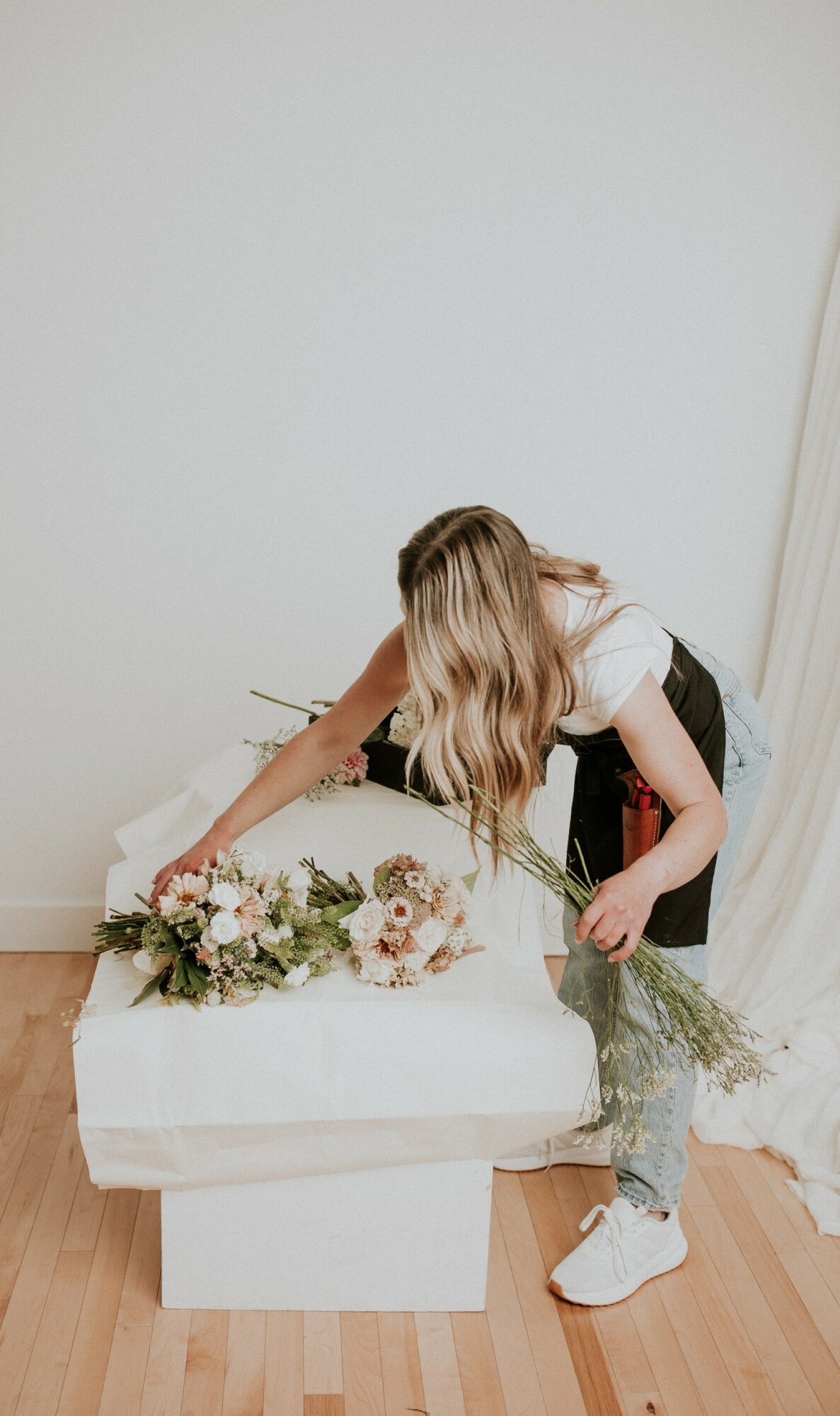 Person arranging a bouquet of flowers on a white table, with a plain white wall background, wooden floor, and casual clothing.