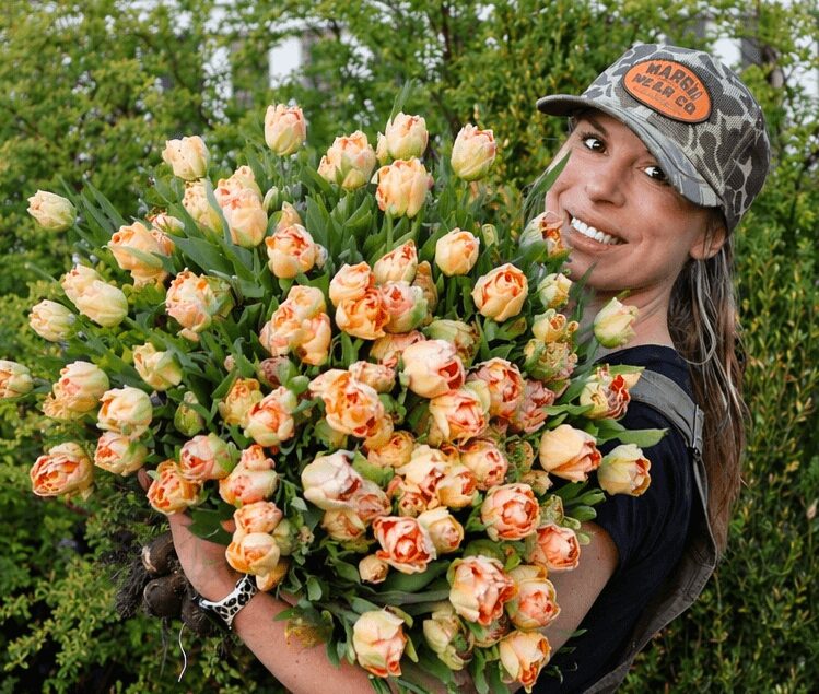 Woman holding a large bouquet of peach and pink roses outdoors, smiling, wearing a camouflage cap and black shirt.