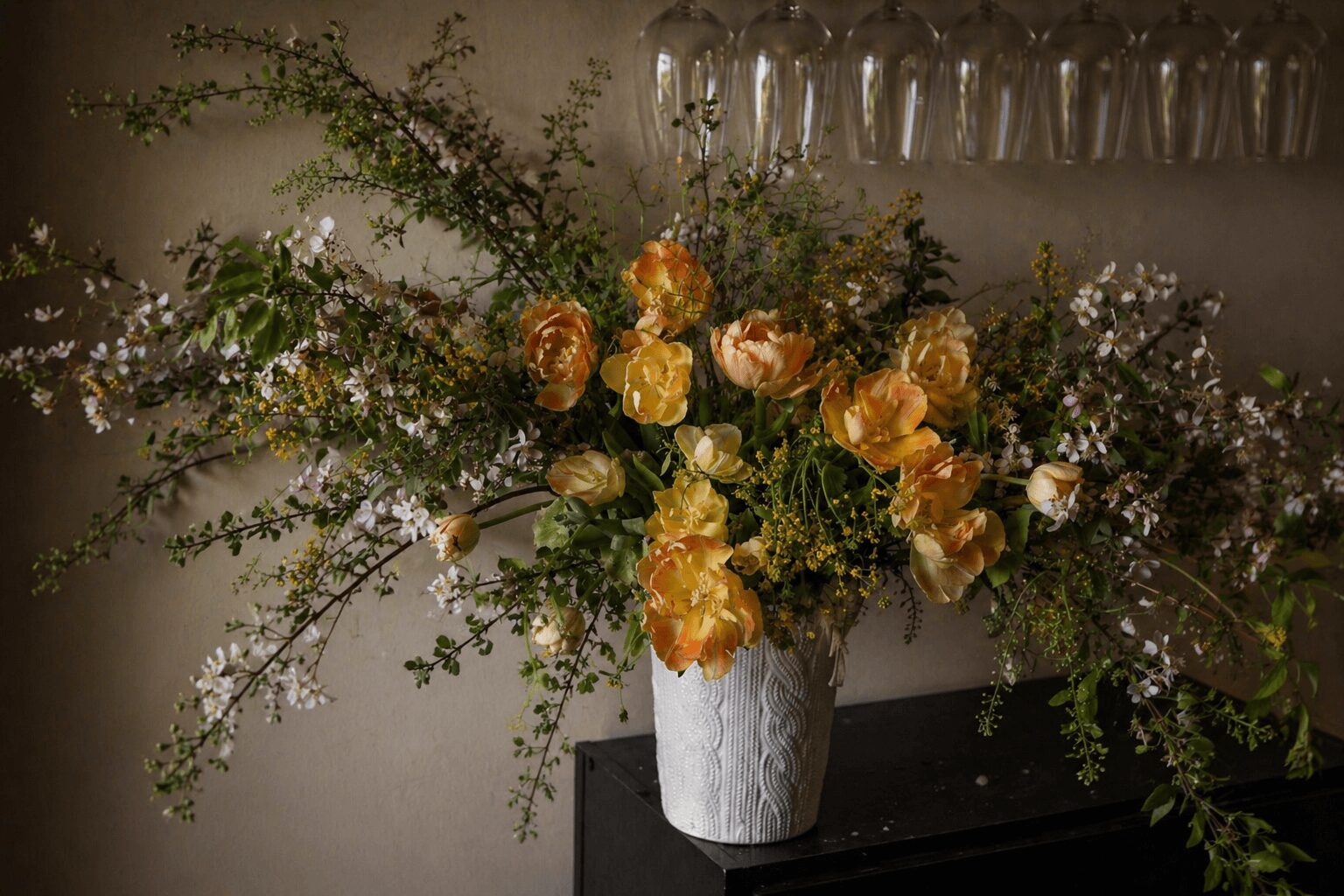 Flower arrangement with yellow, orange, and pink blooms in a white vase on a dark surface, with a glass rack in the background.