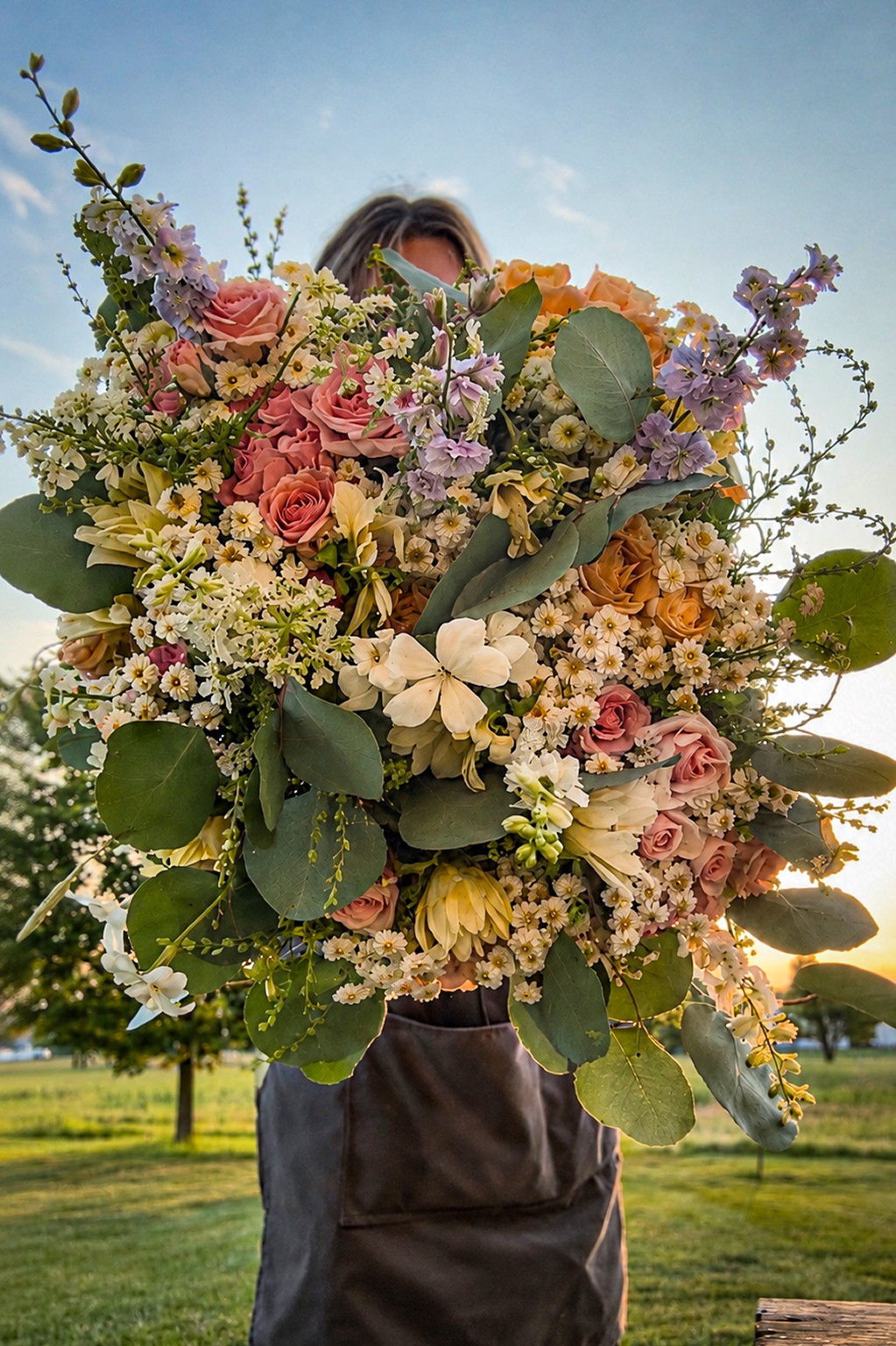 Person holding a large bouquet of mixed flowers outdoors, with a blue sky and trees in the background.
