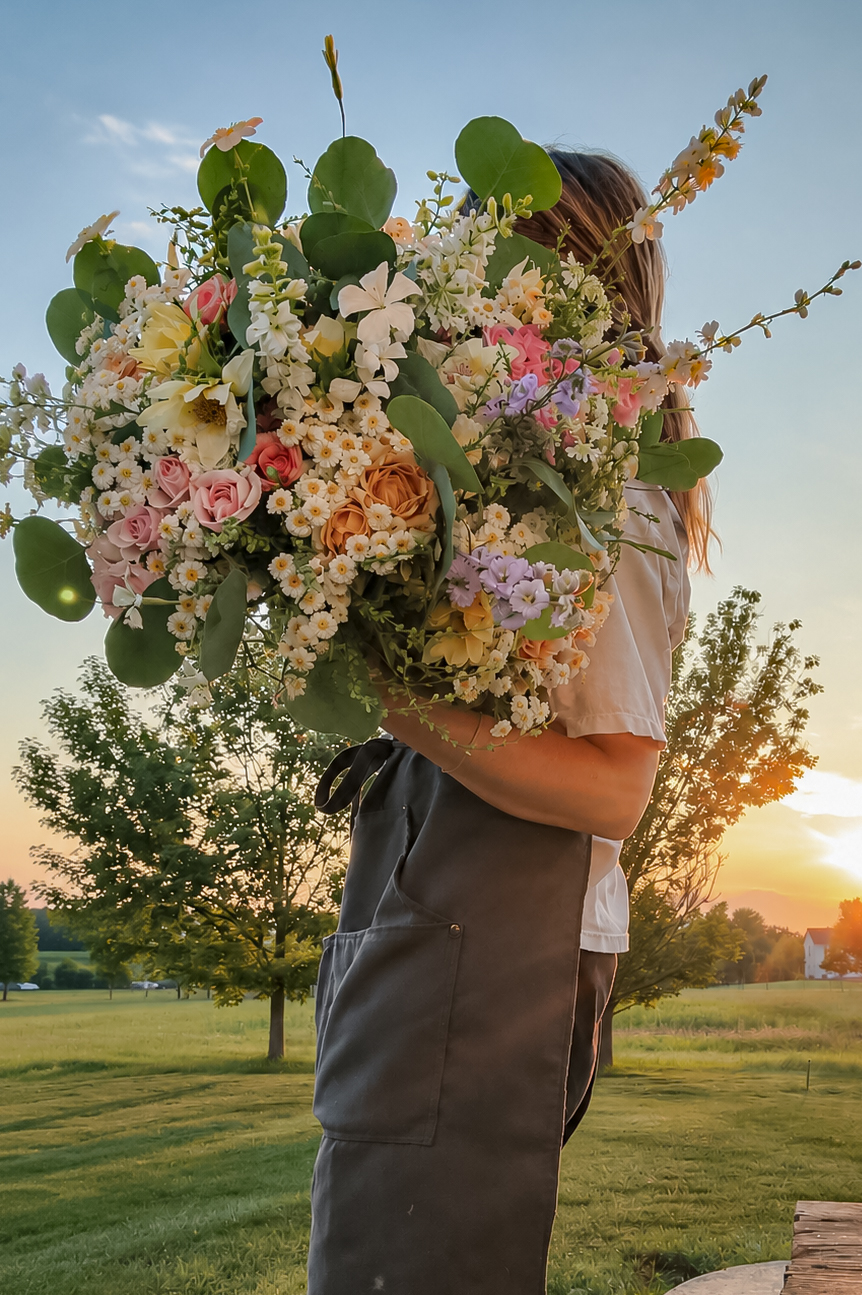 Person holding a large colorful flower bouquet outdoors during sunset, with trees and a grassy field in the background.