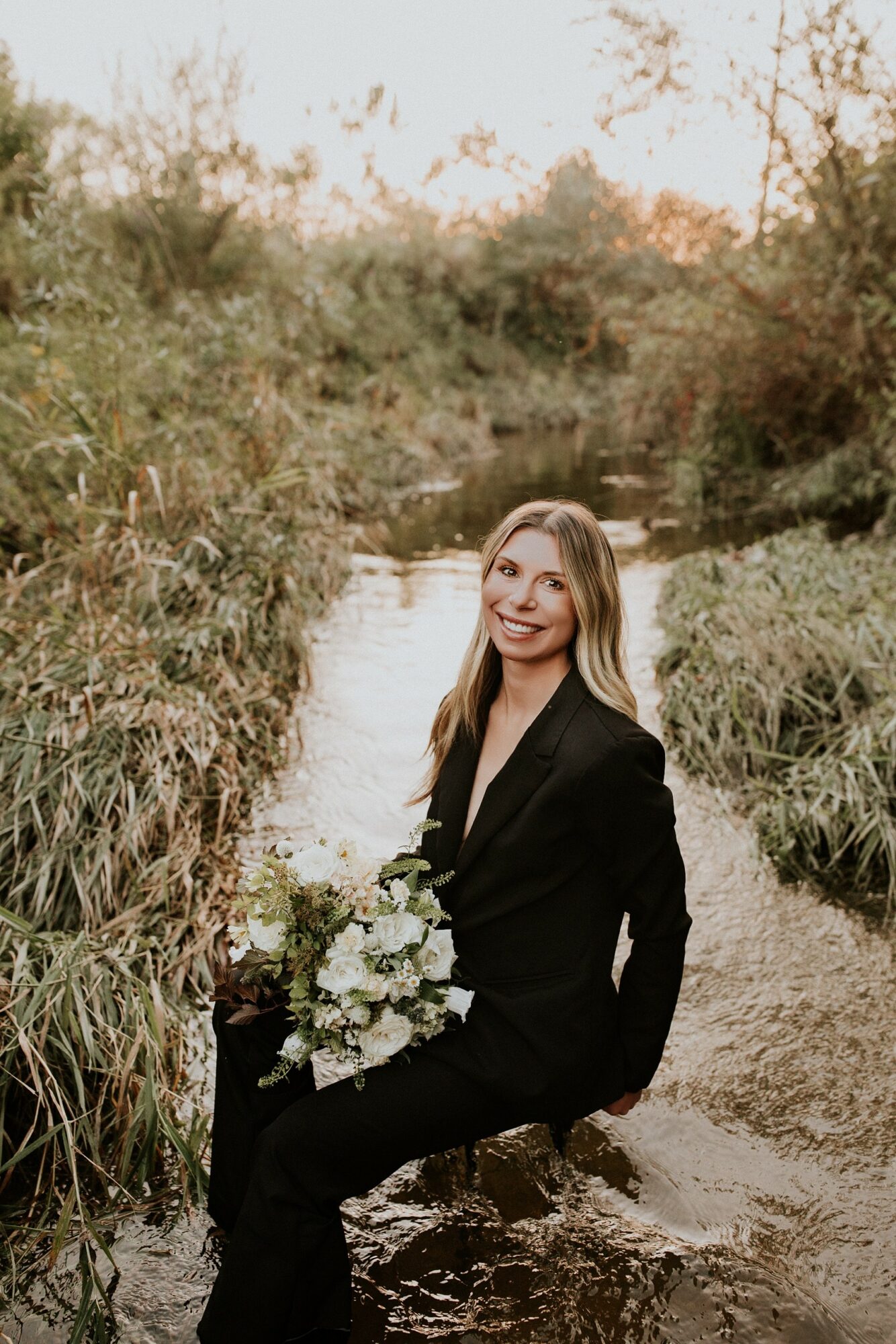 Woman in black suit sitting on a rock by a stream, holding a bouquet, outdoors with trees and sky in background.