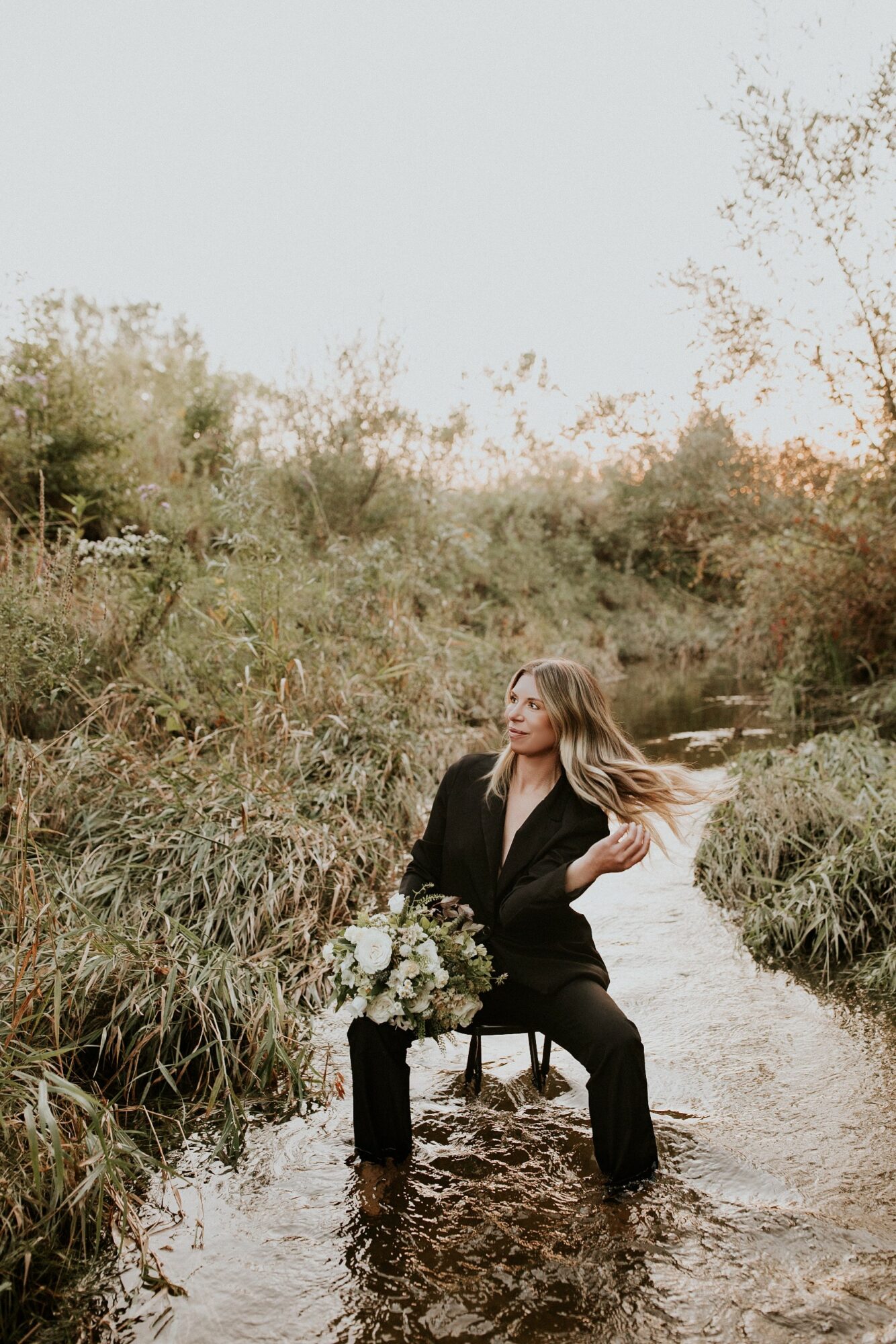 Woman in black pants and blazer sitting in a shallow stream surrounded by tall grass and trees, with flowing hair.