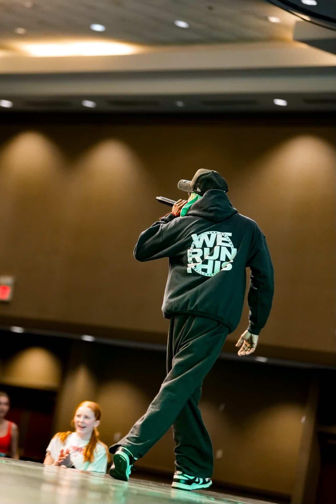 Person in black hoodie and cap walking on stage with two children watching, in a theater or auditorium setting.
