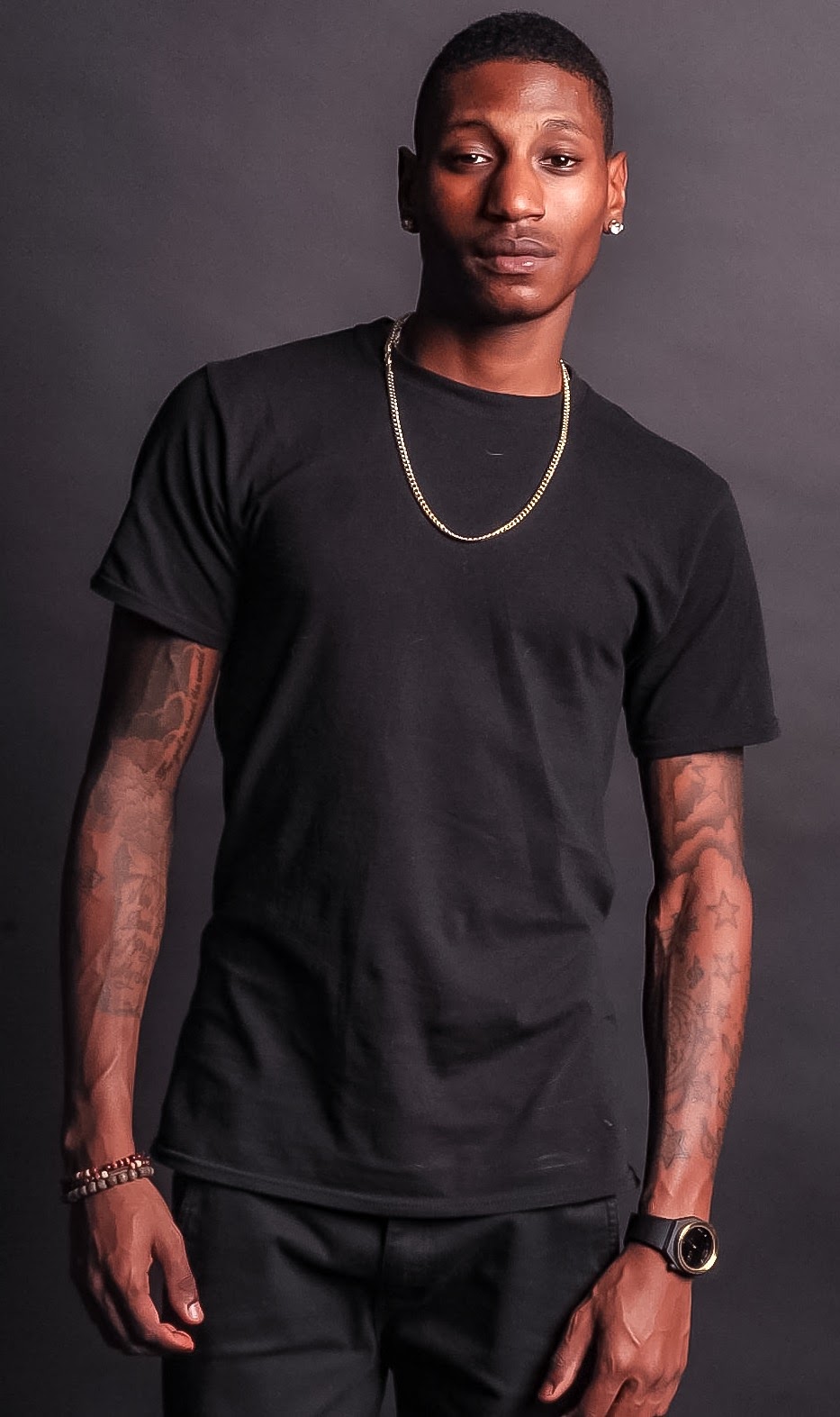 Young person with short hair, wearing a black t-shirt, gold chain, bracelet, and watch, standing against a dark background.