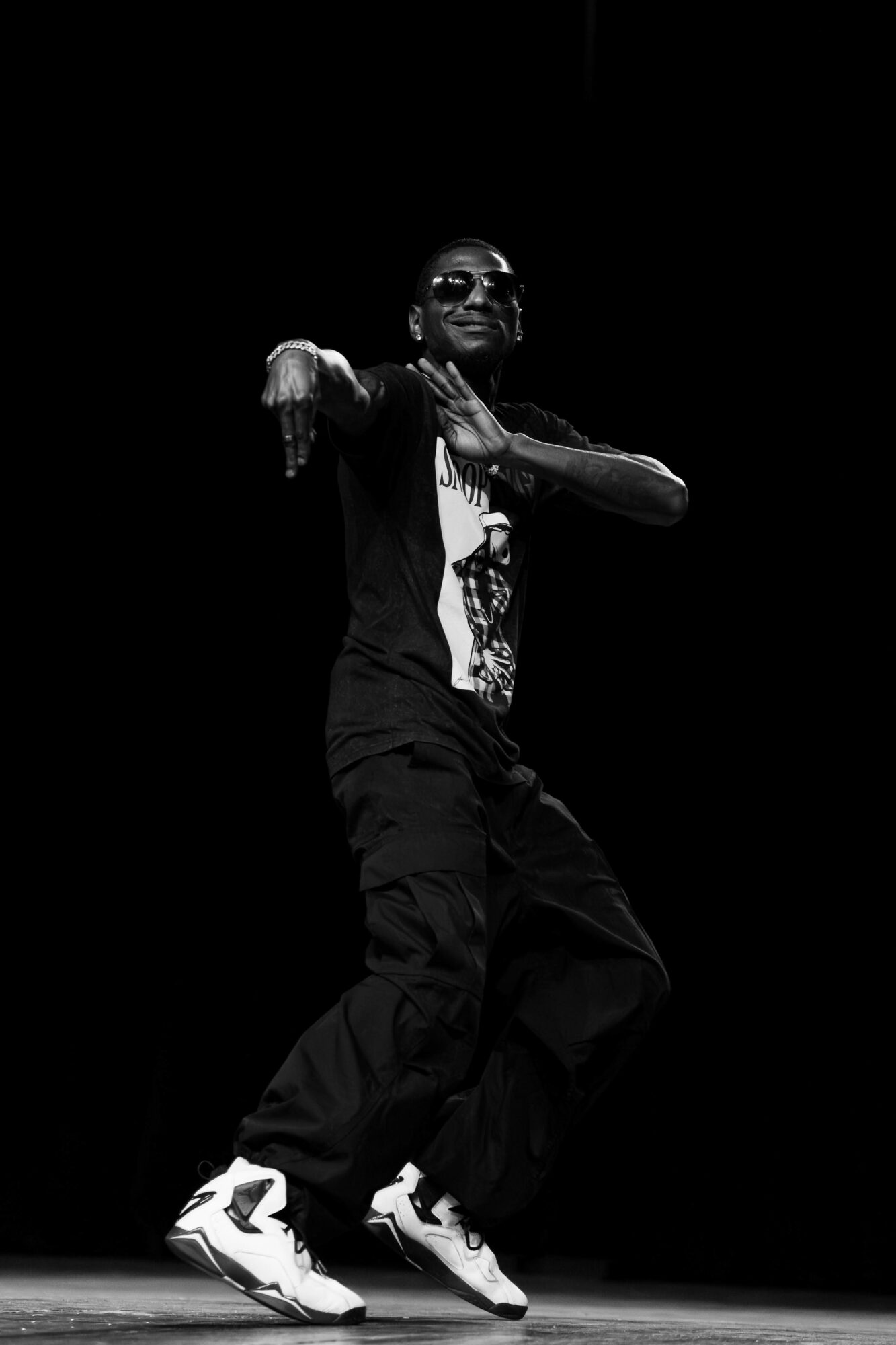 Person dancing on stage, wearing sunglasses, black clothing, and sneakers, with a dark background.