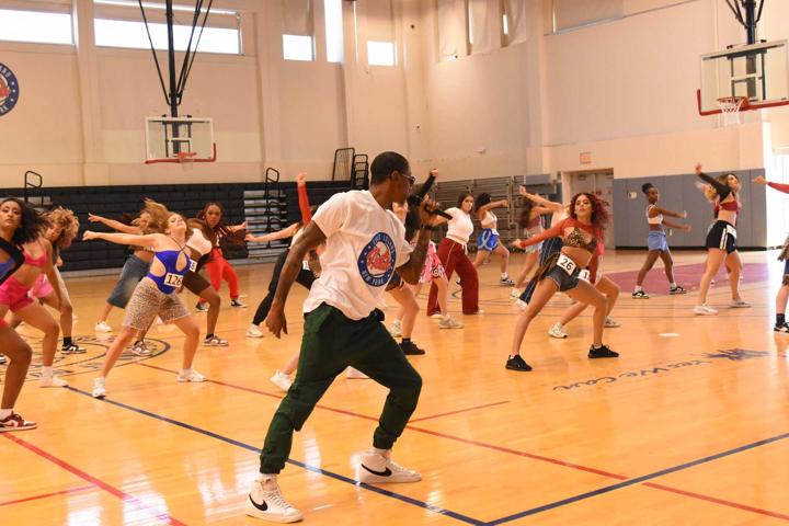 Group of people dancing in a gymnasium with basketball hoops, some with arms extended, wearing casual clothing.
