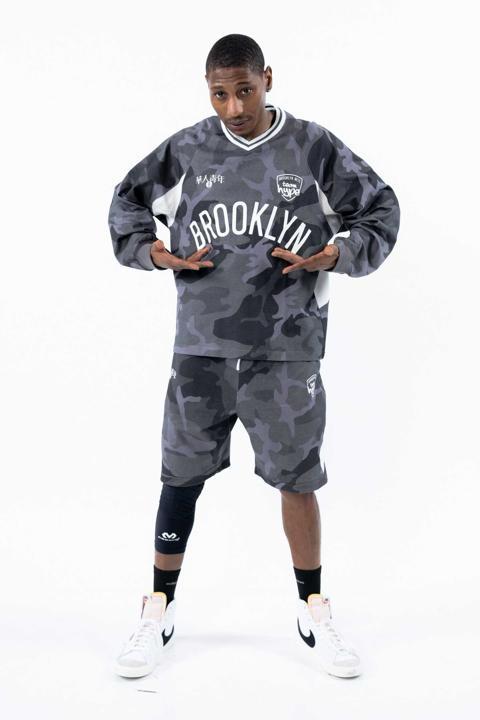 Young person in camouflage sportswear holding a Brooklyn jersey, standing with legs apart, white sneakers, and a neutral background.