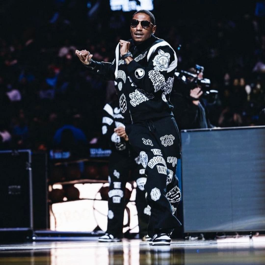 Person performing on stage with a microphone, wearing sunglasses and a black and white patterned outfit, in a dark setting.