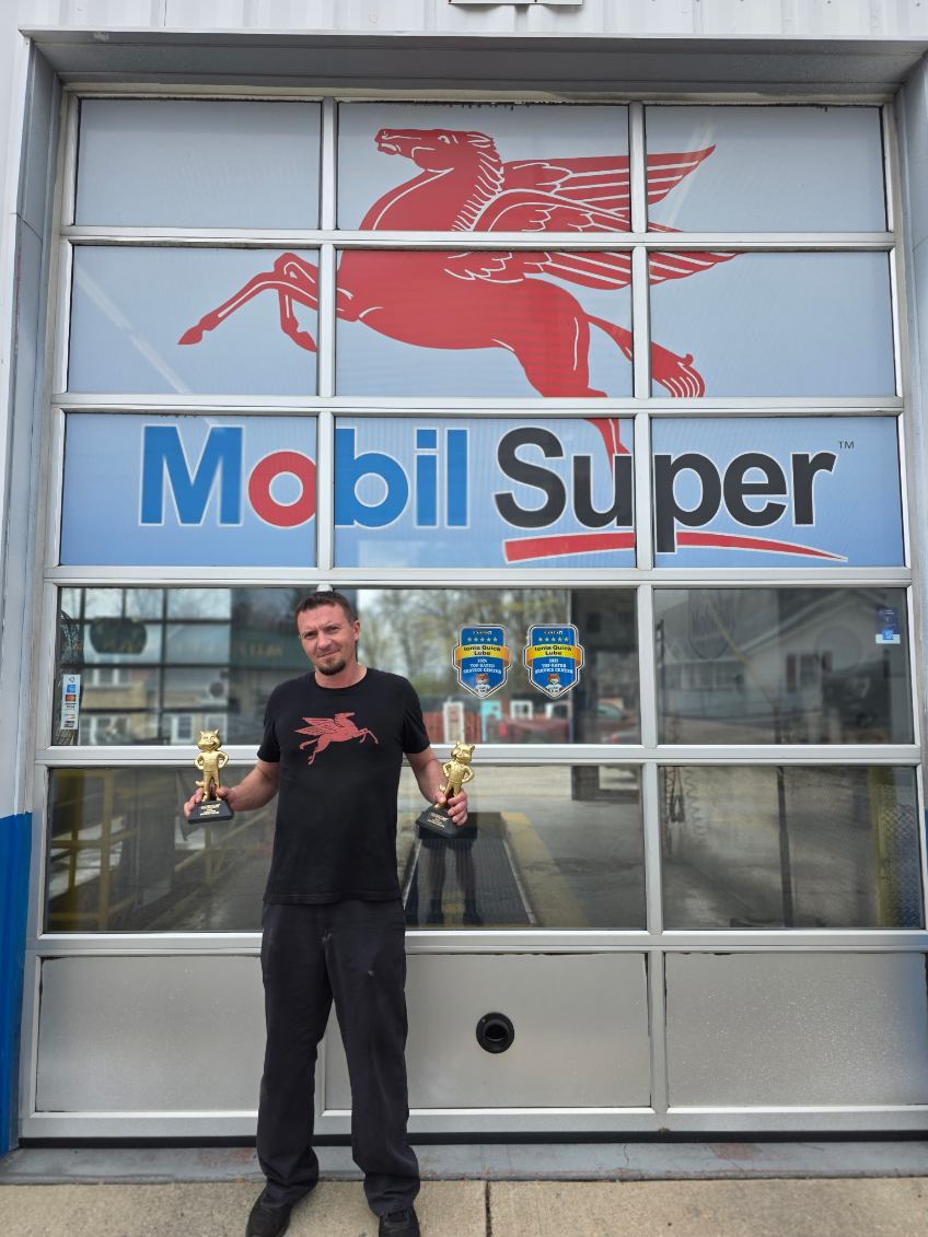 Man holding trophies standing in front of a large window with Mobil Super logo and a red Pegasus symbol.