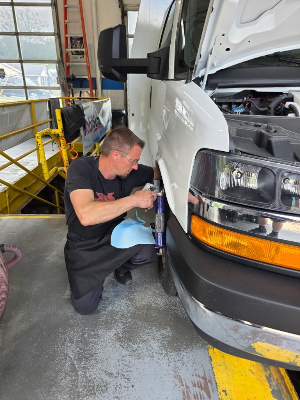 Man kneeling beside a white vehicle, working on the front side in a workshop with yellow safety barriers.