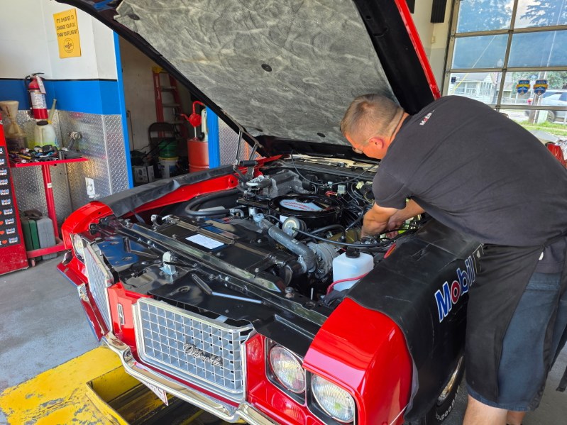 Person working on car engine inside garage, hood open, red and black vehicle, tools nearby, large windows in background.
