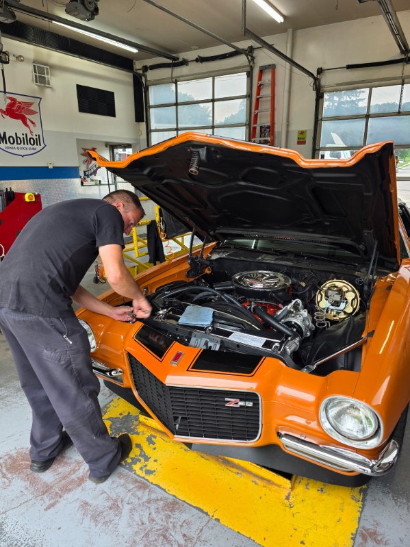 Person working on an orange car with open hood in garage, tools and equipment visible, garage windows in background.