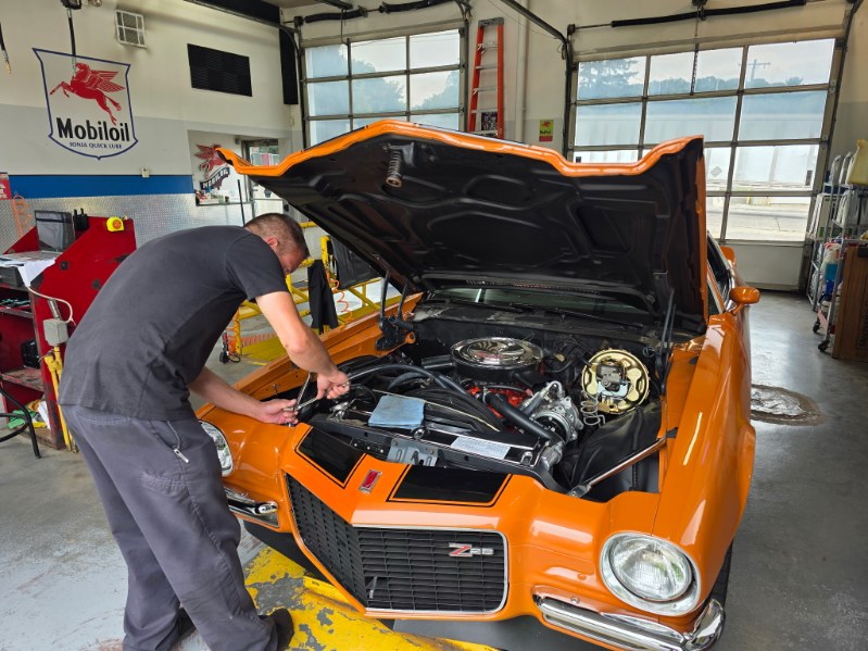 Mechanic working on an orange car engine inside a garage with large windows and tools around.