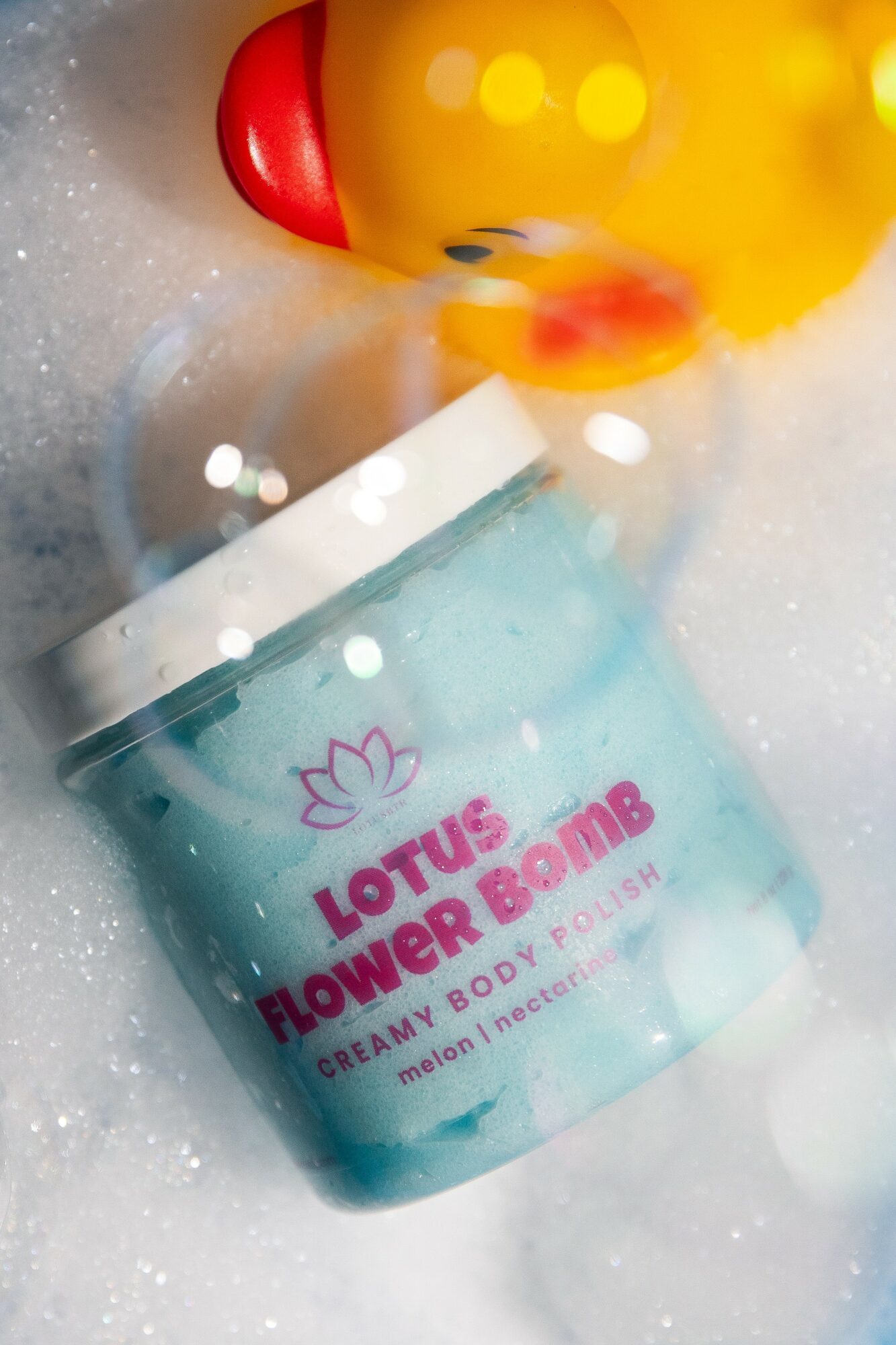 Container of Lotus Flower & Bomb bath product with a rubber duck and bubbles around it.