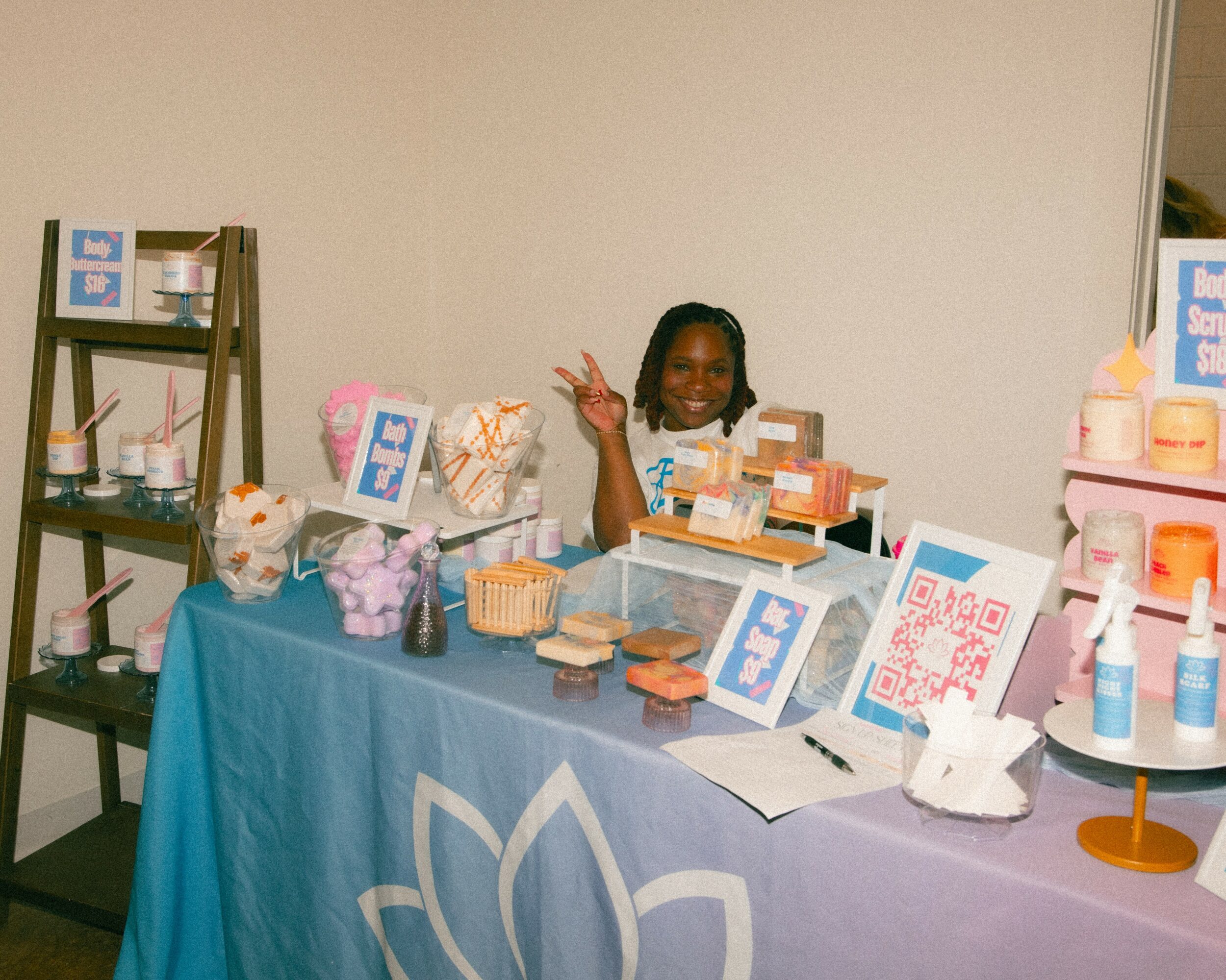 Child smiling behind a table with skincare products and signs, waving hand, in a room with plain wall.