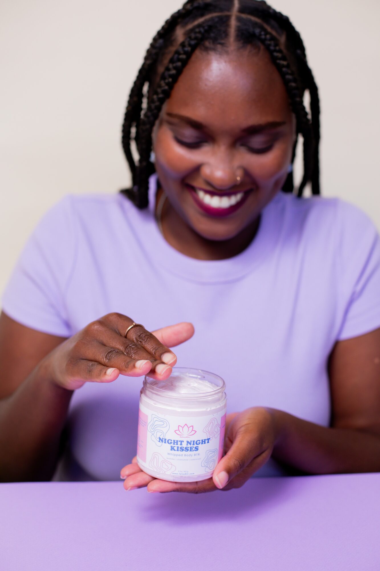 Smiling woman with braided hair opening a jar of skincare cream, wearing a light purple shirt.