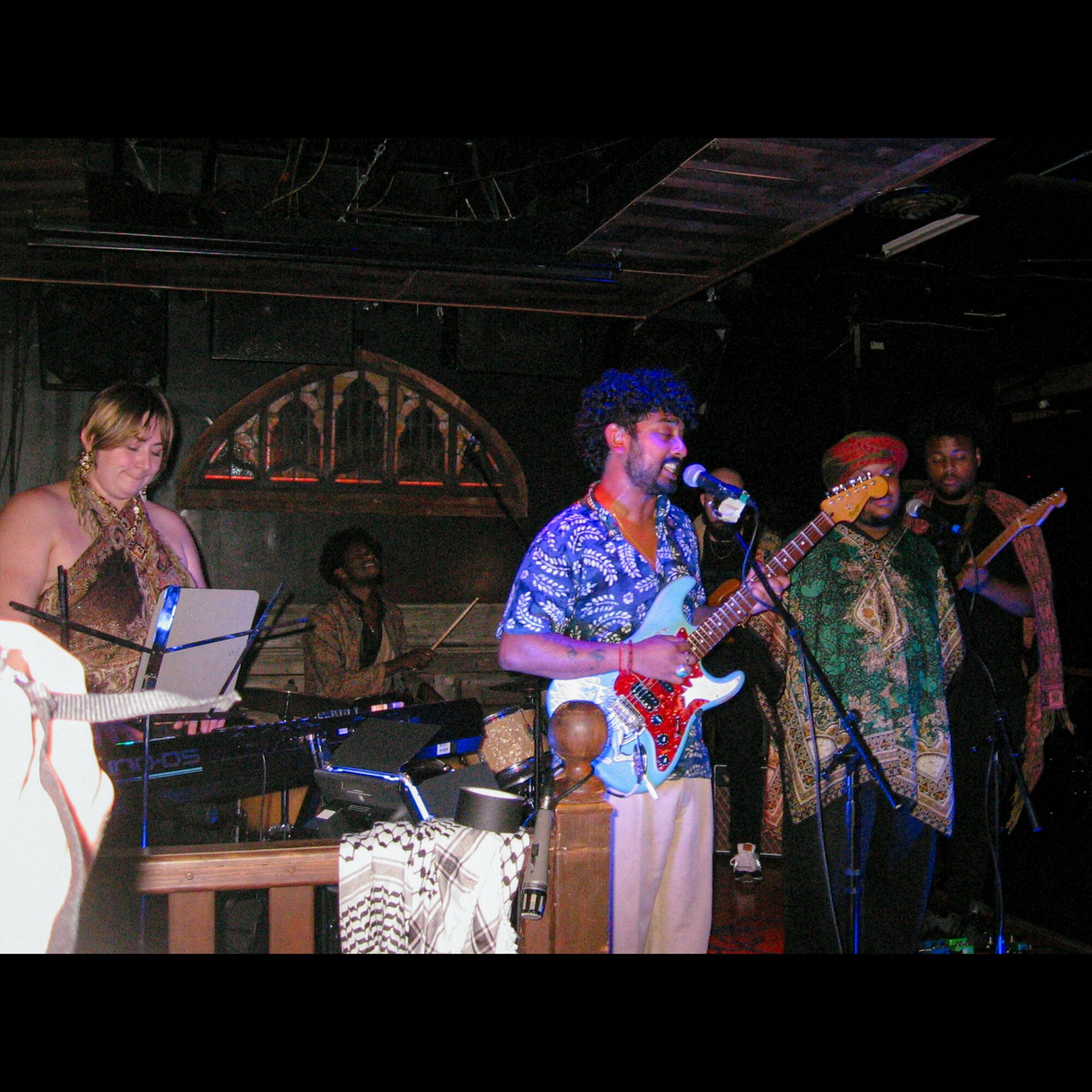 Group of musicians performing on stage with instruments, including a keyboard, guitar, and drums, in a dimly lit venue.