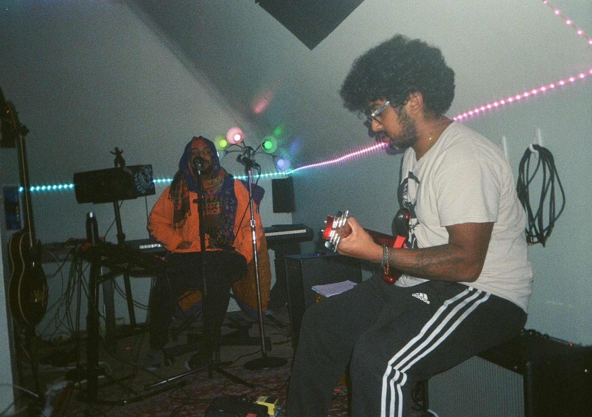 Two people in a music studio, one playing guitar and the other sitting with a microphone, colorful lights in the background.