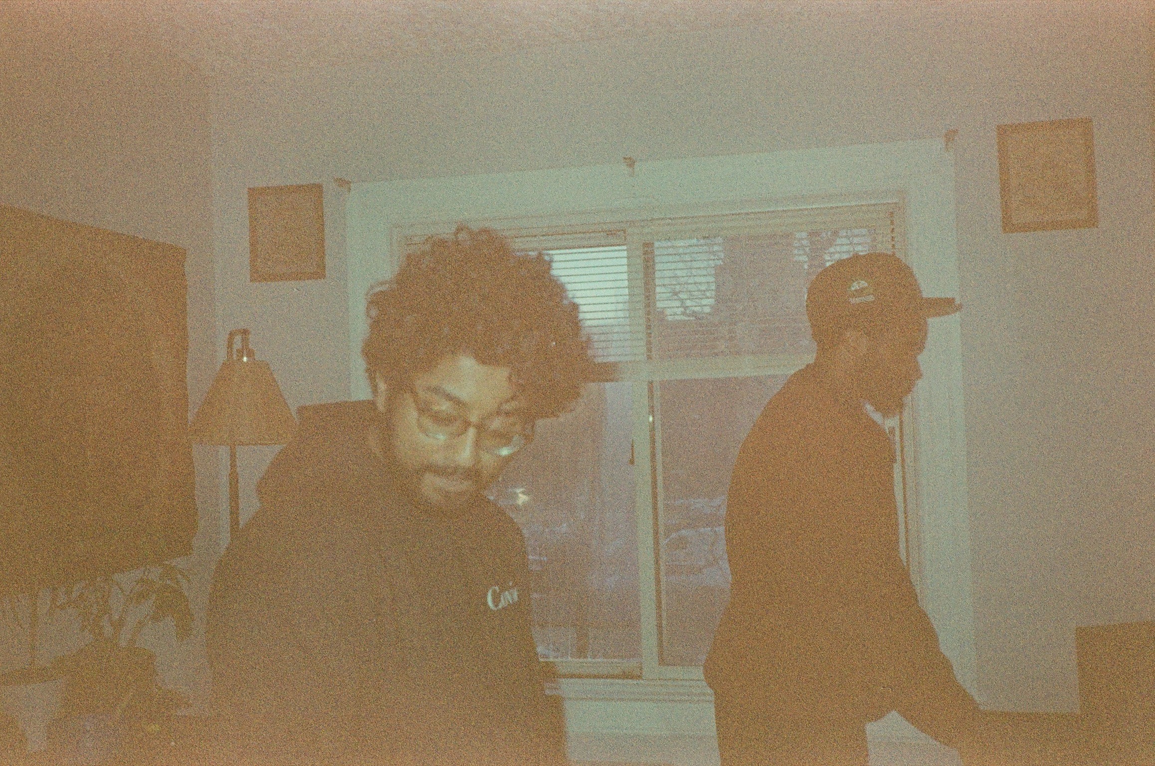 Two people in a room with a window, one with curly hair and glasses, the other wearing a cap, facing away.