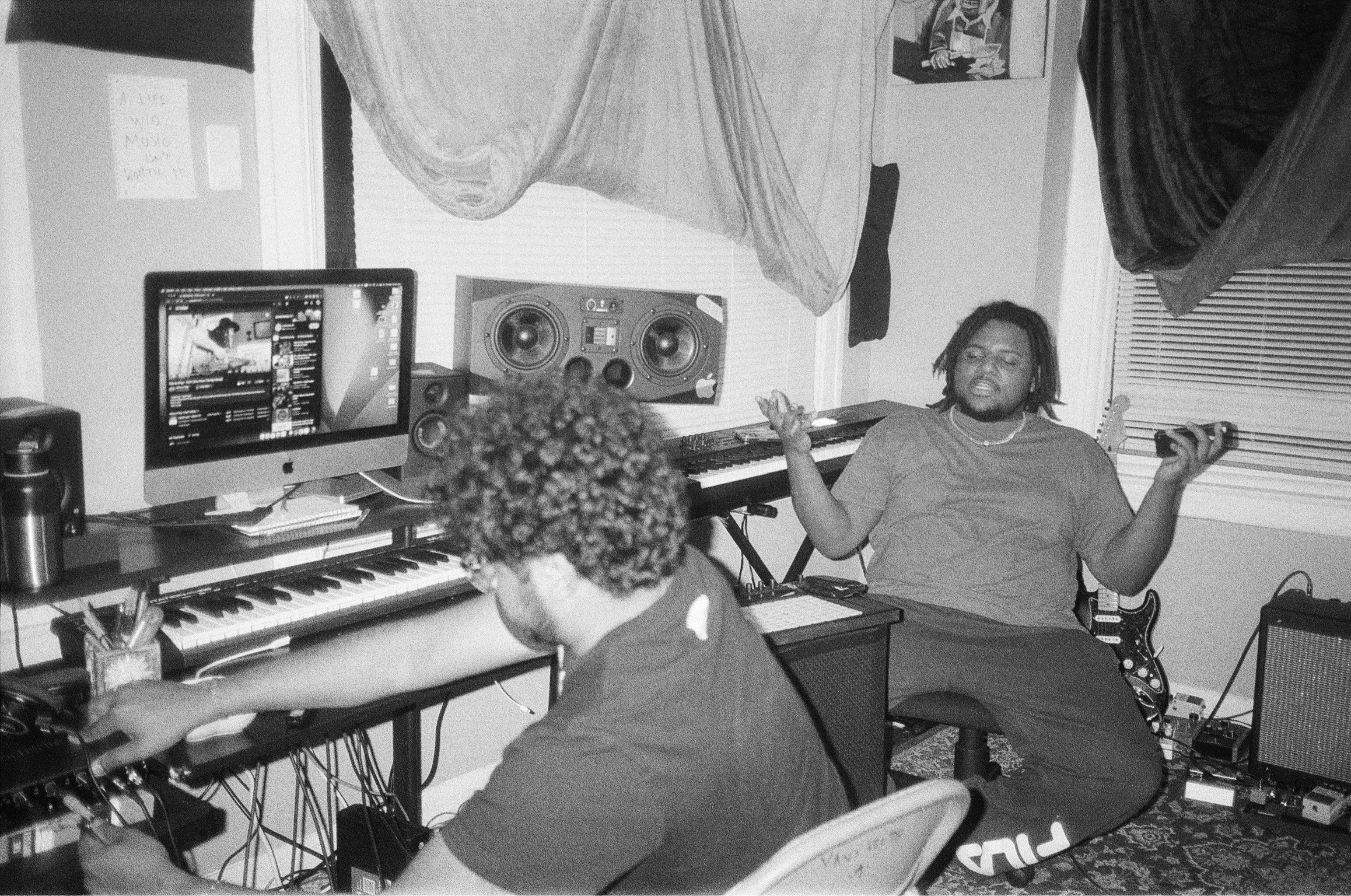 Two people in a music studio with computers, keyboard, and speakers; one person with curly hair working at the keyboard, another with glasses sitting on the floor with arms outstretched.