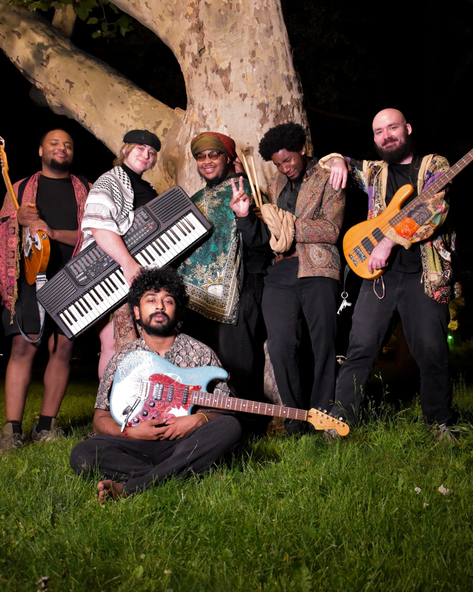 Group of six people outdoors at night, holding musical instruments, standing near a large tree, smiling and posing.