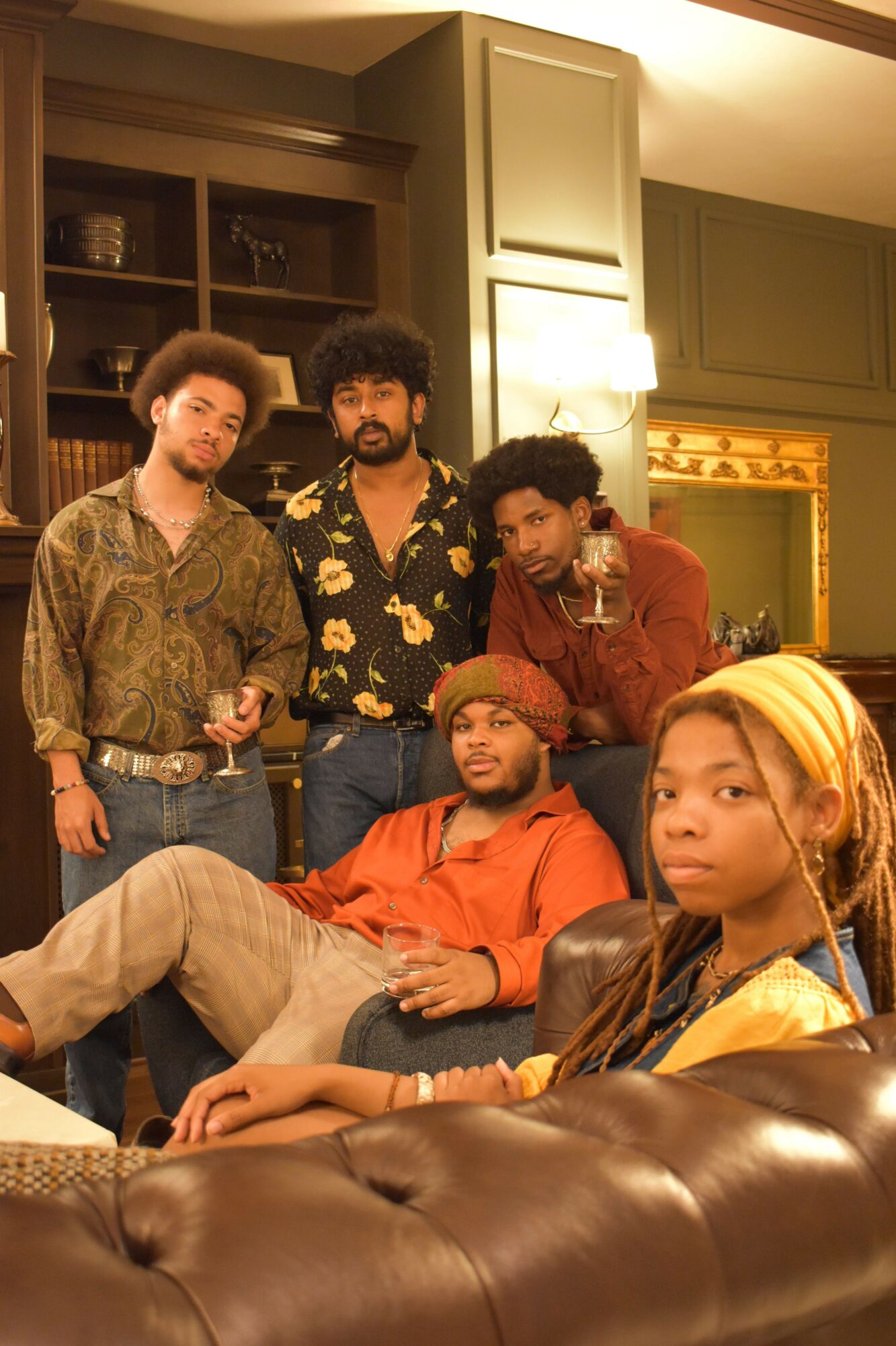 Group of five people sitting on a couch in a living room, some holding drinks, with a bookshelf and mirror behind them.