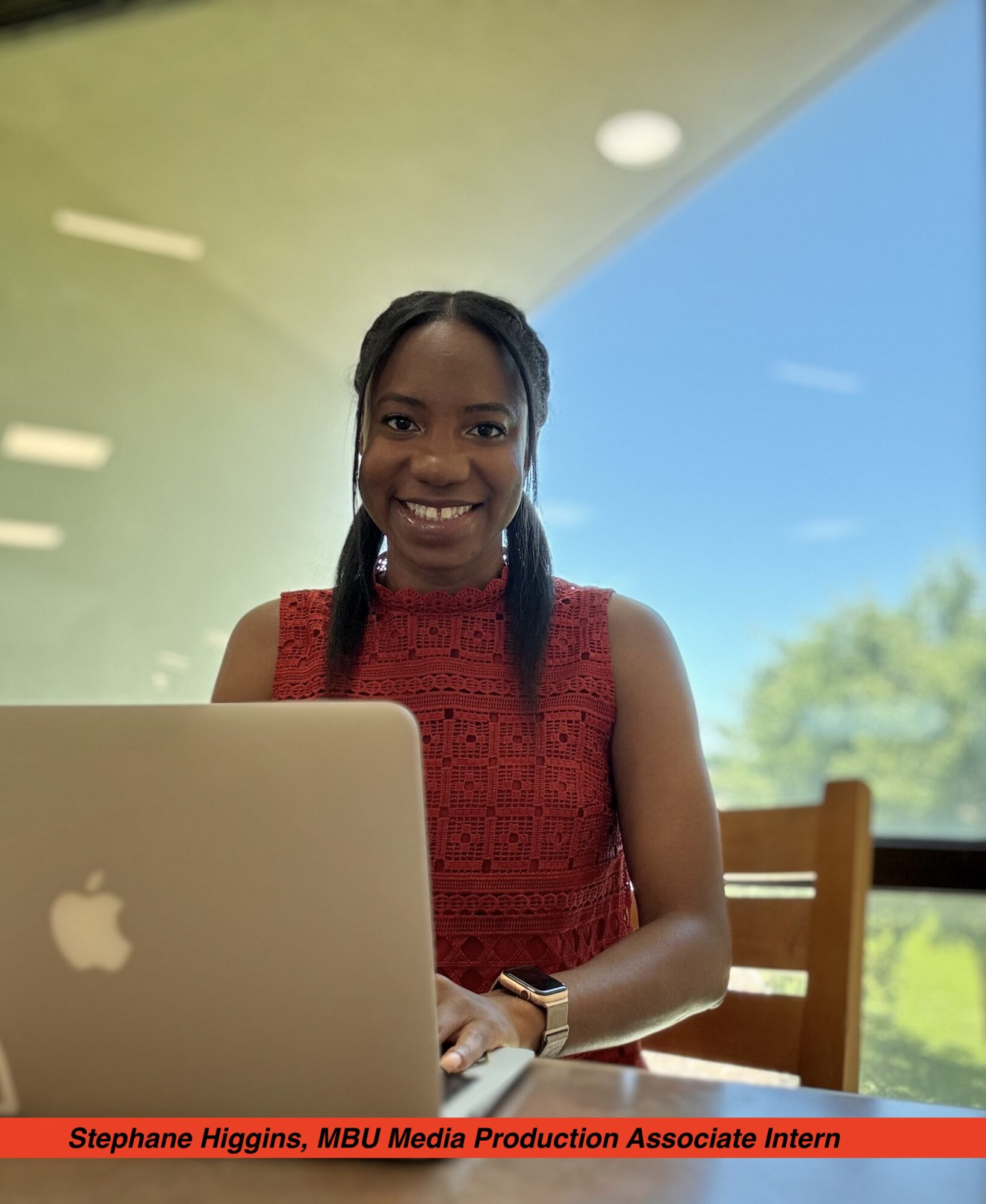 Smiling woman sitting at a table with a laptop, window showing trees and sky behind her.