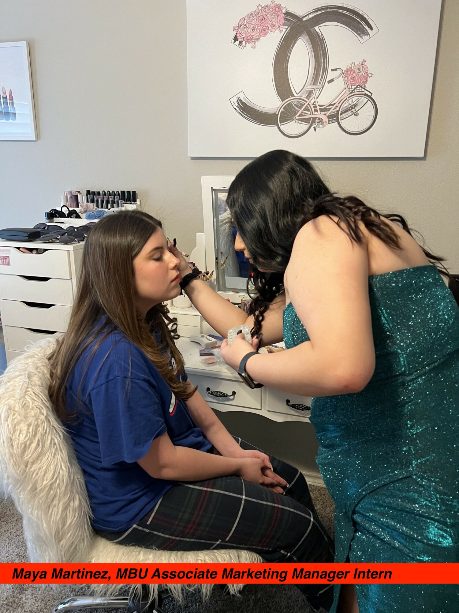 A woman in a teal dress applies makeup to a seated young woman with long brown hair, in a room with artwork on the wall.